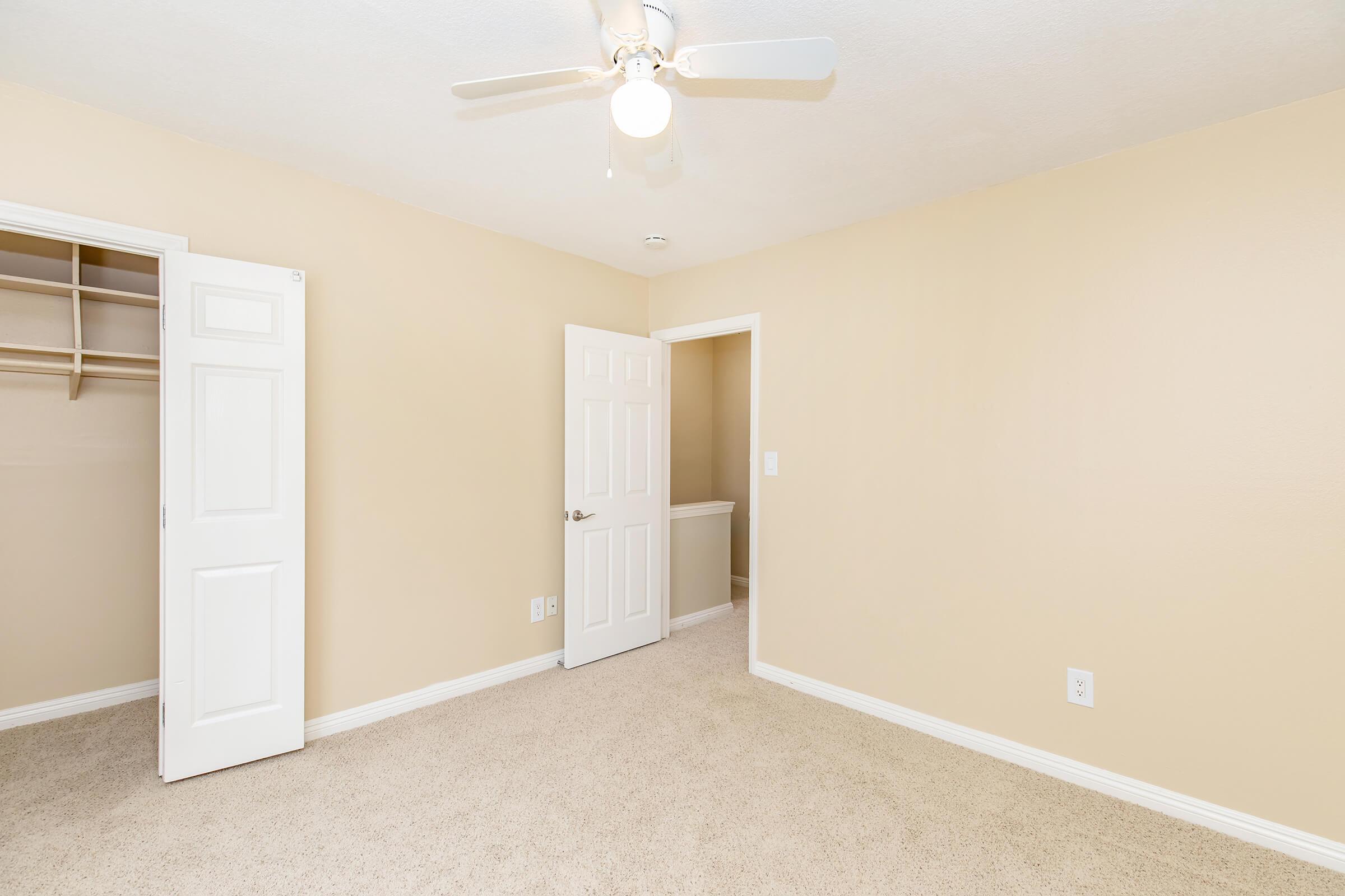 A well-lit, empty bedroom featuring beige walls and carpet. There is a ceiling fan with three blades, a closet with sliding doors on one side, and a door leading to another room on the opposite side. The overall space has a clean and minimalist appearance, ready for decoration.
