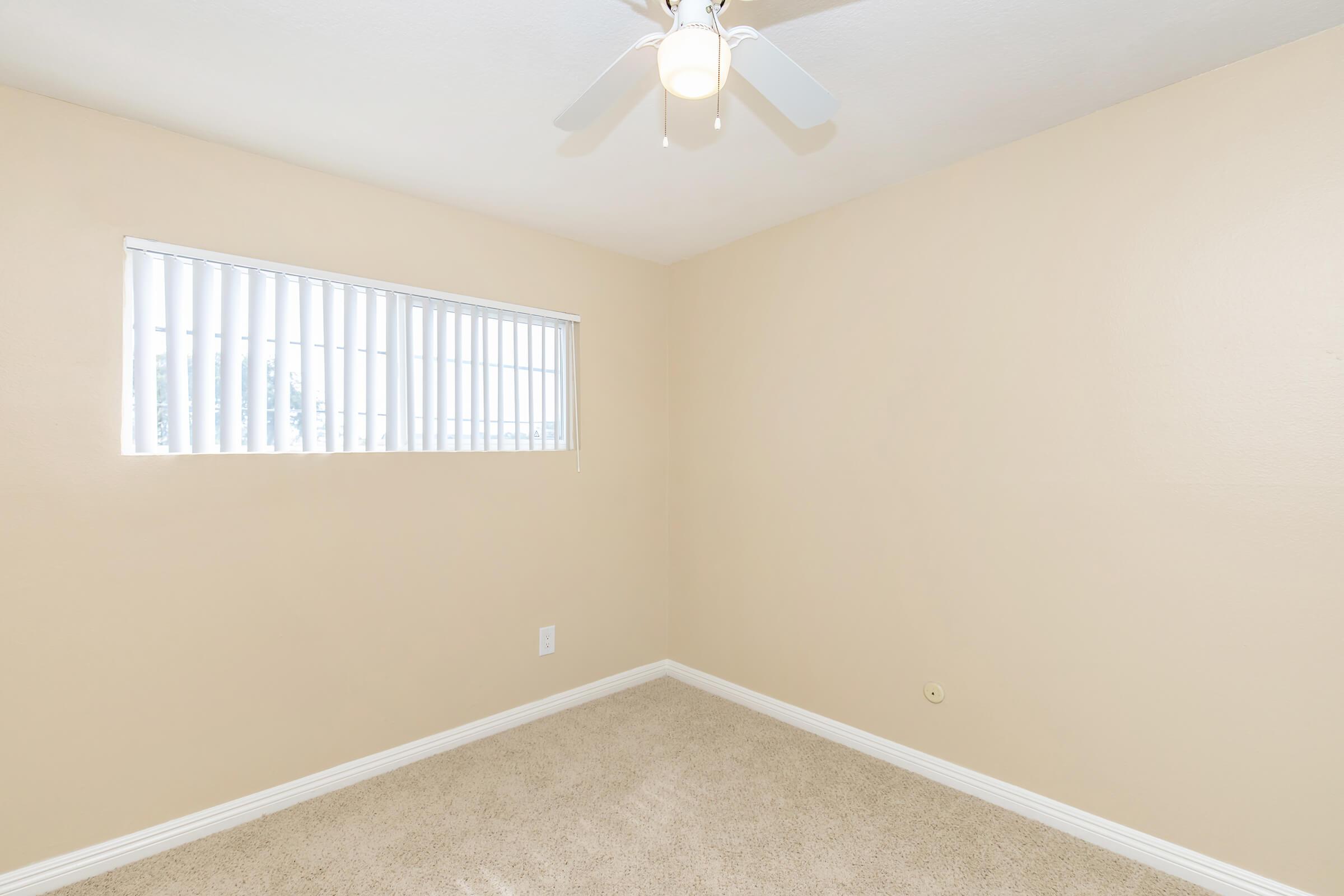 A vacant room with light beige walls and a ceiling fan. There is a window with white vertical blinds letting in natural light. The room features soft, carpeted flooring and has minimal furnishings, creating a clean and simple aesthetic.