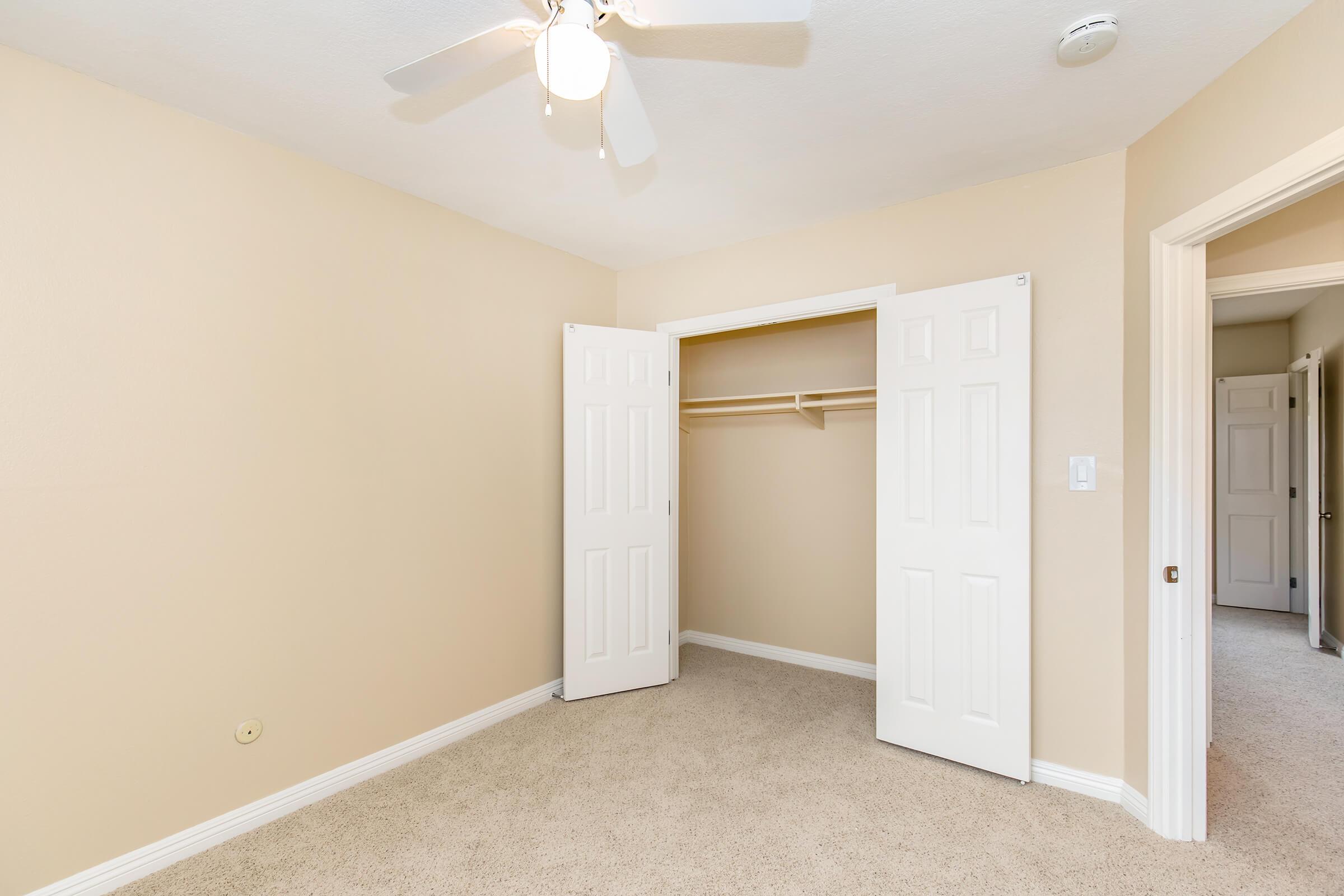 A light-colored bedroom with beige walls and plush carpet. The room features a ceiling fan and an open closet with double doors on the left. The doorway to another room is visible on the right. The overall atmosphere is clean and well-lit, creating a welcoming space.