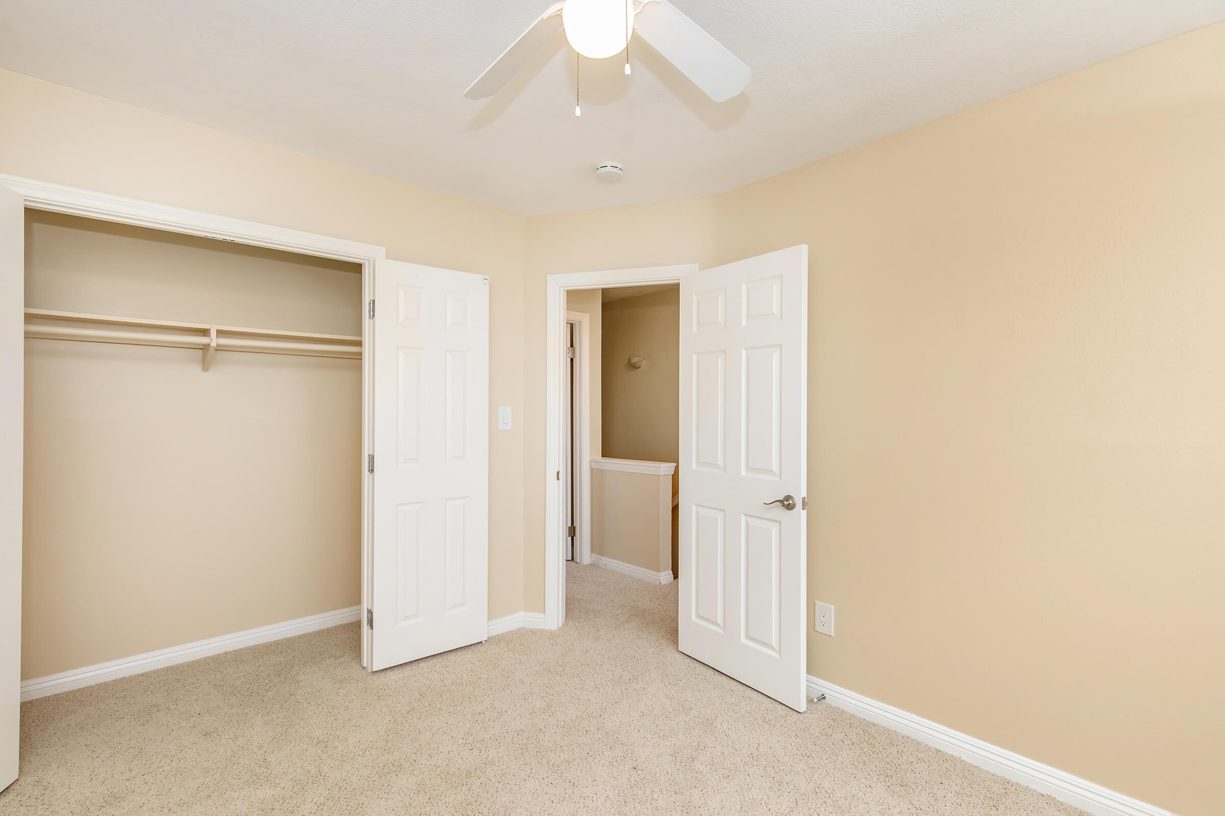A light-filled room with beige walls and carpet, featuring two open doors leading to a closet and hallway. A ceiling fan is mounted above, enhancing the airy feel of the space. The neutral color palette creates a warm and inviting atmosphere.