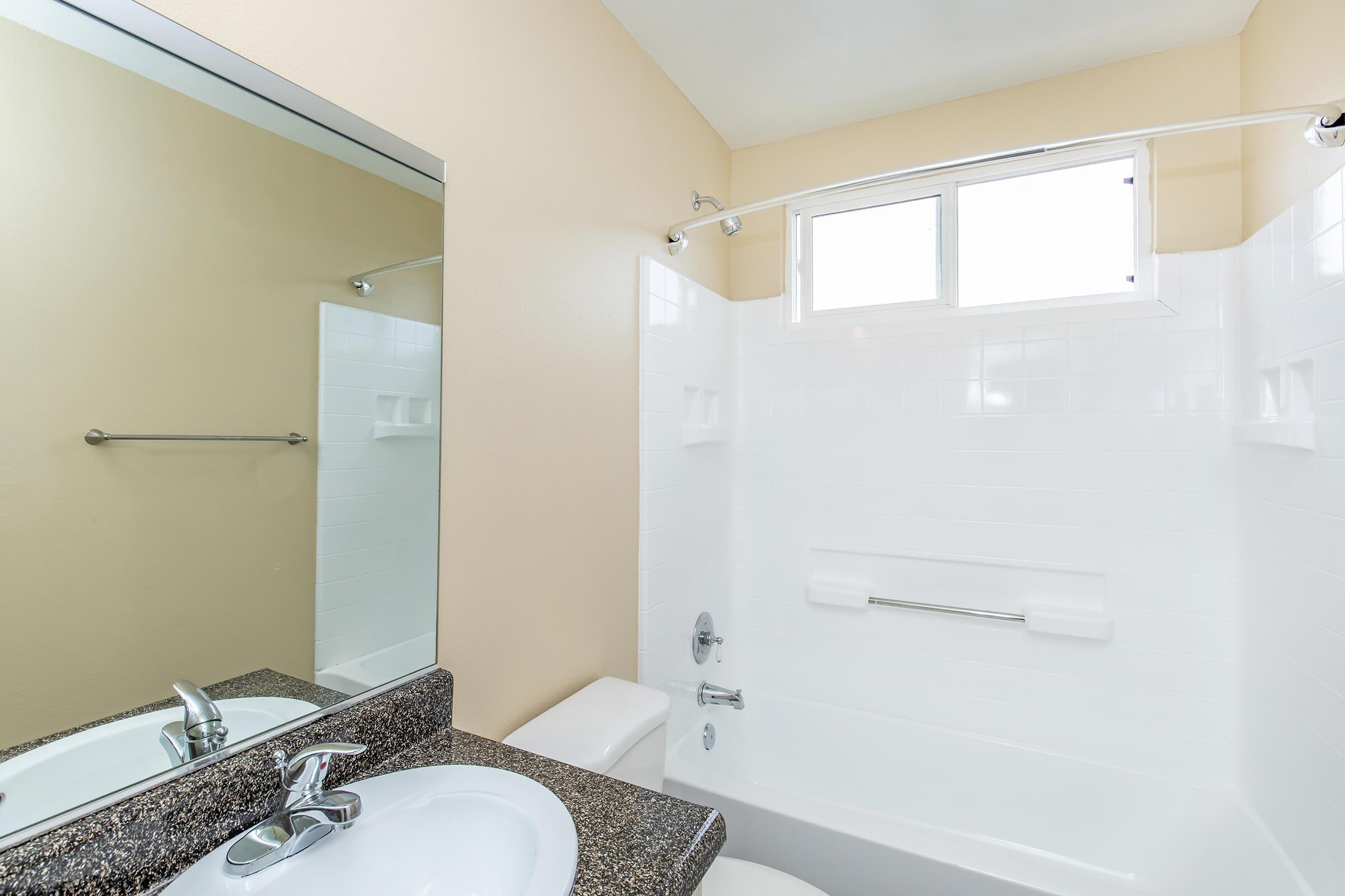 A clean and modern bathroom featuring a white bathtub and shower combination, a granite countertop sink with a mirror above, and a window providing natural light. The walls are painted a neutral color, and there are towel racks and built-in shelves visible.