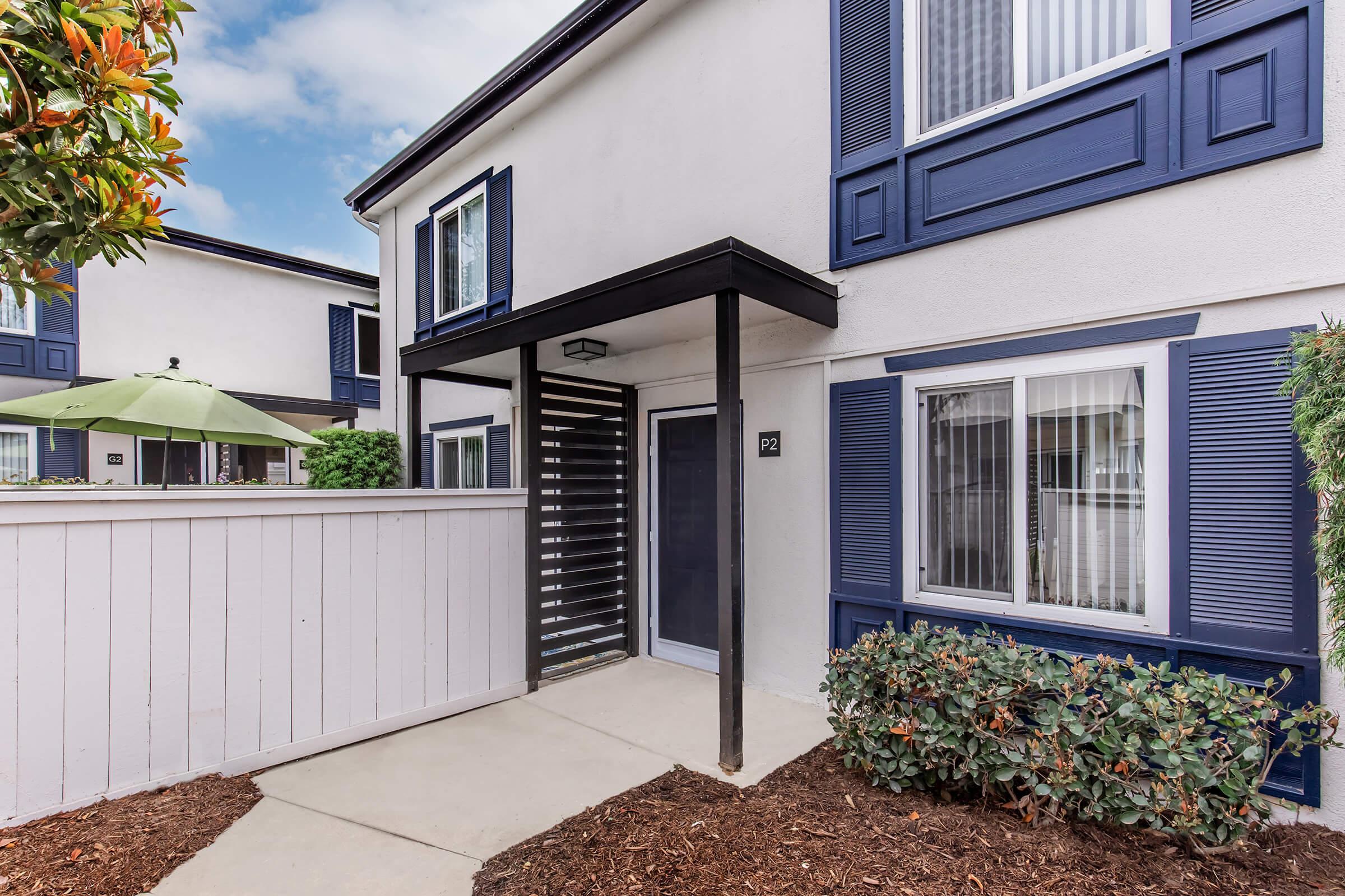A two-story residential building featuring a white exterior with blue accents. The entrance is marked by a modern canopy and a decorative lattice screen. Surrounding the entrance are neatly trimmed shrubs and a small pathway leading to the door, with an umbrella and greenery visible nearby.