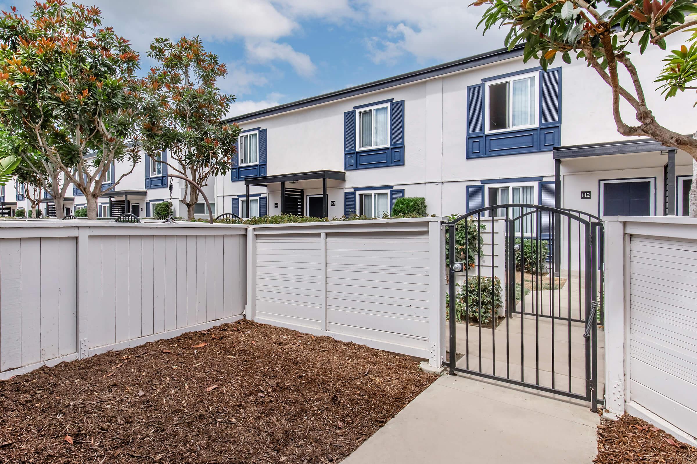 A serene outdoor area featuring a gated entrance surrounded by a white fence. The path leads to a row of modern townhouses with blue shutters and lush greenery. A few trees add a touch of nature, and the sky is partly cloudy, enhancing the tranquil atmosphere.