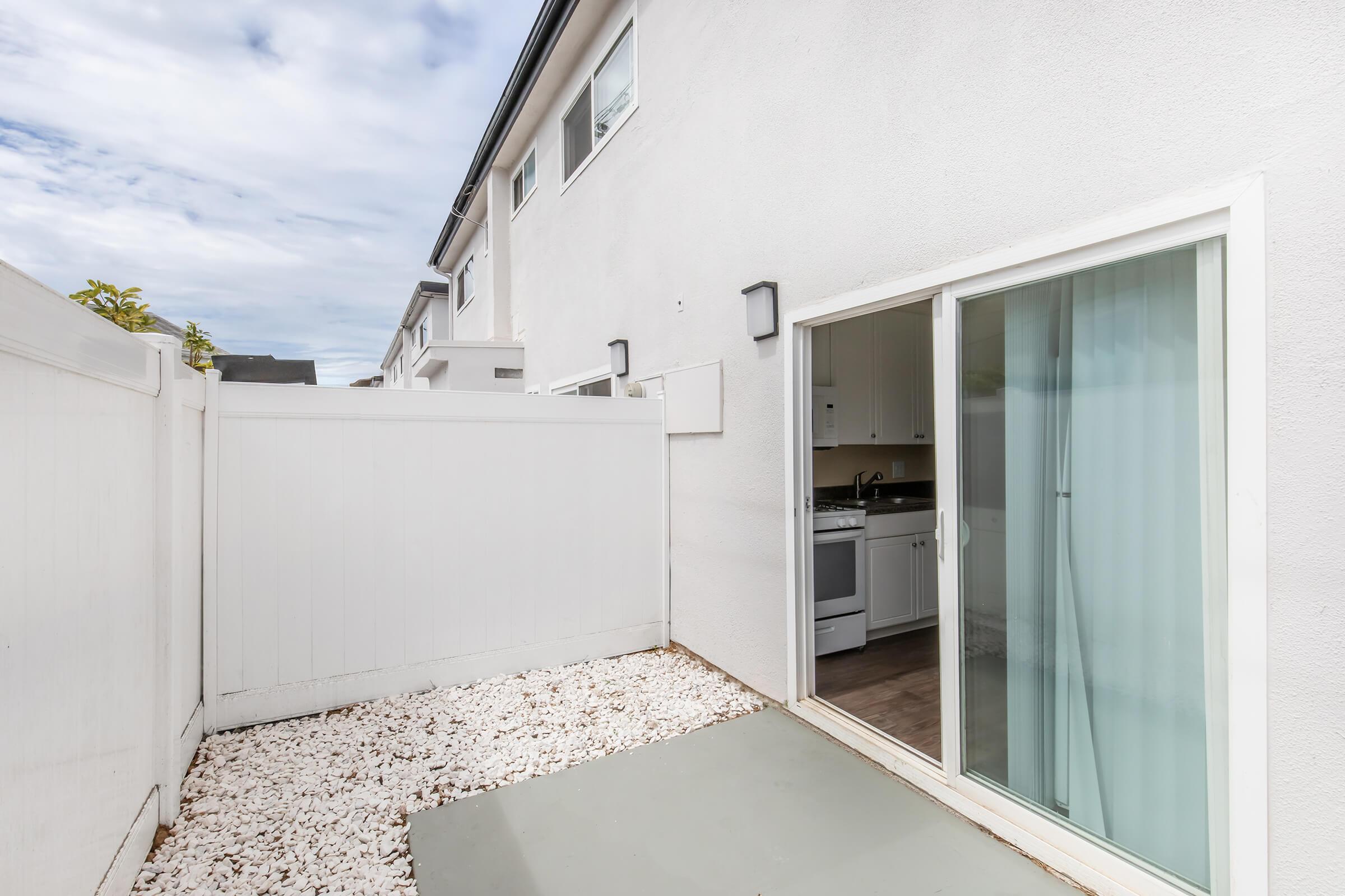 A small outdoor patio area with white gravel and a sliding glass door leading to a kitchen. The patio is enclosed by tall white fences, and there are neighboring buildings visible in the background under a cloudy sky.