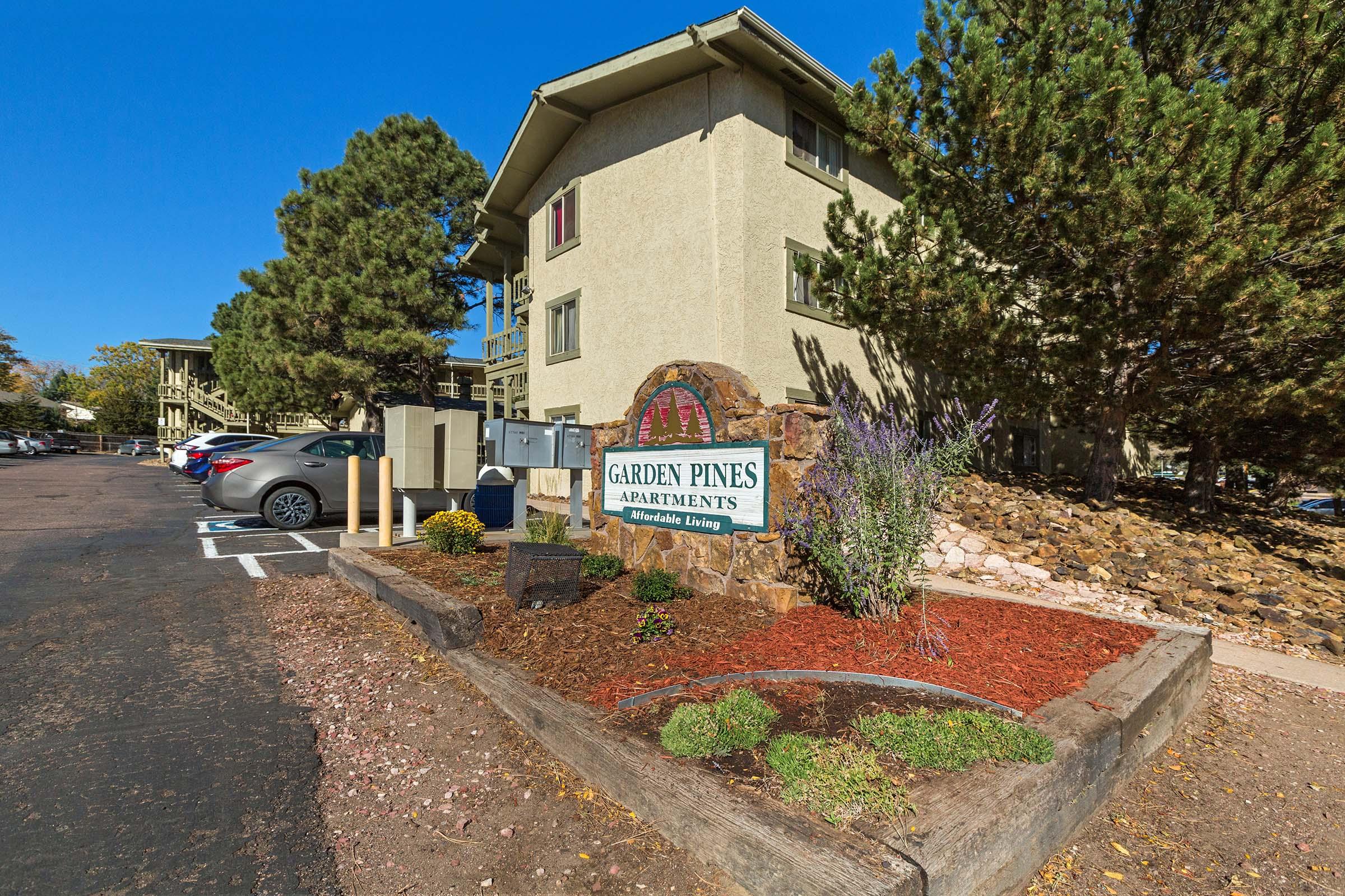 Sign for Garden Pines Apartments in a landscaped area with gravel and plants. Nearby, there are parked cars and several trees. In the background, a multistory building is visible under a clear blue sky. The setting suggests a residential community with a focus on affordable living.