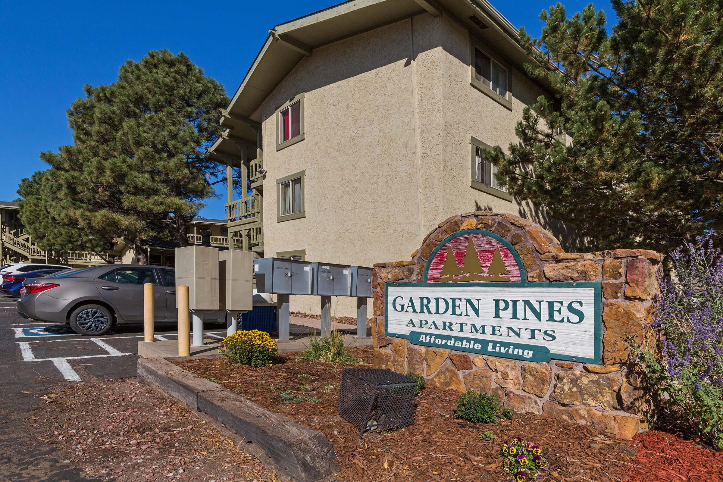The image shows a multi-story residential building with the sign "Garden Pines Apartments" prominently displayed in front. There are mailboxes nearby, parking spaces, and landscaped areas with flowers and shrubs. The setting appears to be well-maintained and offers affordable living options.