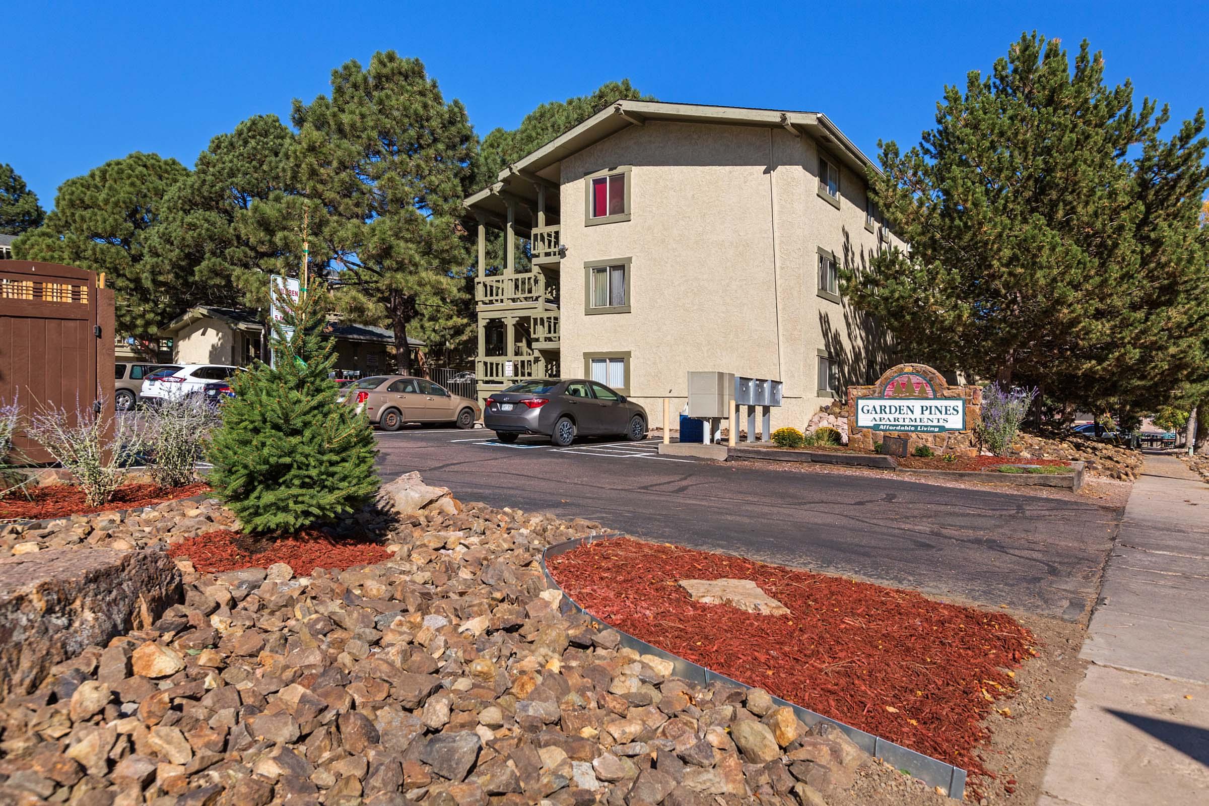 An apartment building with a light-colored exterior, surrounded by trees and landscaping featuring rocks and mulch. A sign reads "Garden Pines." Several parked cars are visible in the lot. The sky is clear and blue, indicating a sunny day.