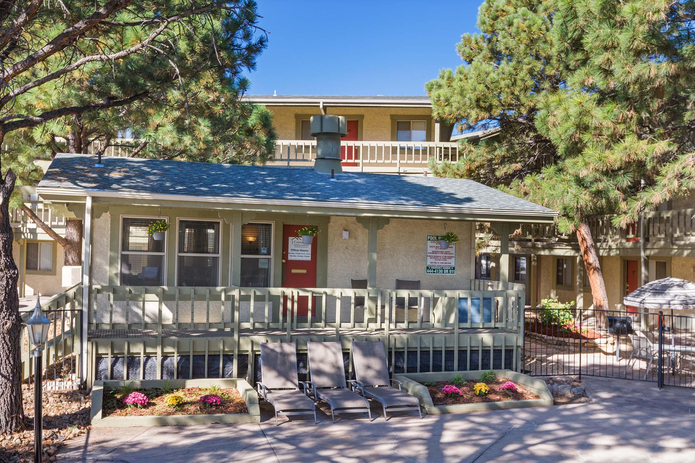 Front view of a charming lodge or apartment building surrounded by tall pine trees. The building features a green porch with simple seating, flowers in planters, and a blue sky above. Sunlight filters through the trees, creating a warm, inviting atmosphere.