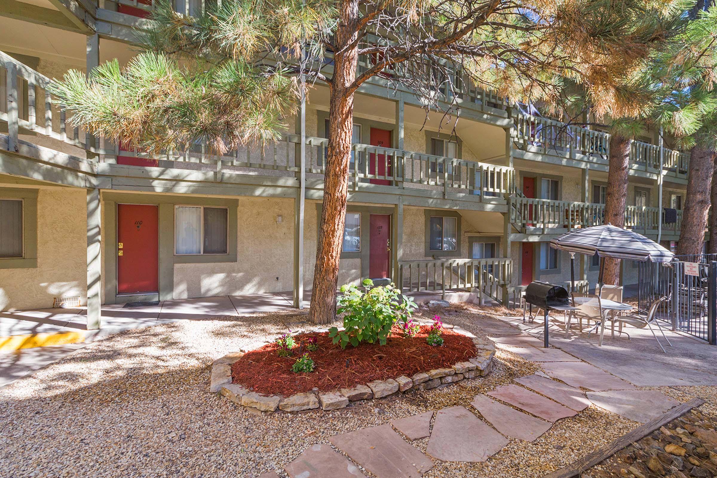 A courtyard view of a multi-story building with balconies, surrounded by pine trees. In the foreground, a landscaped area with colorful flowers and mulch, alongside a stone pathway. A table and chairs are set up under a small shaded patio. Sunlight filters through the trees, creating a serene atmosphere.