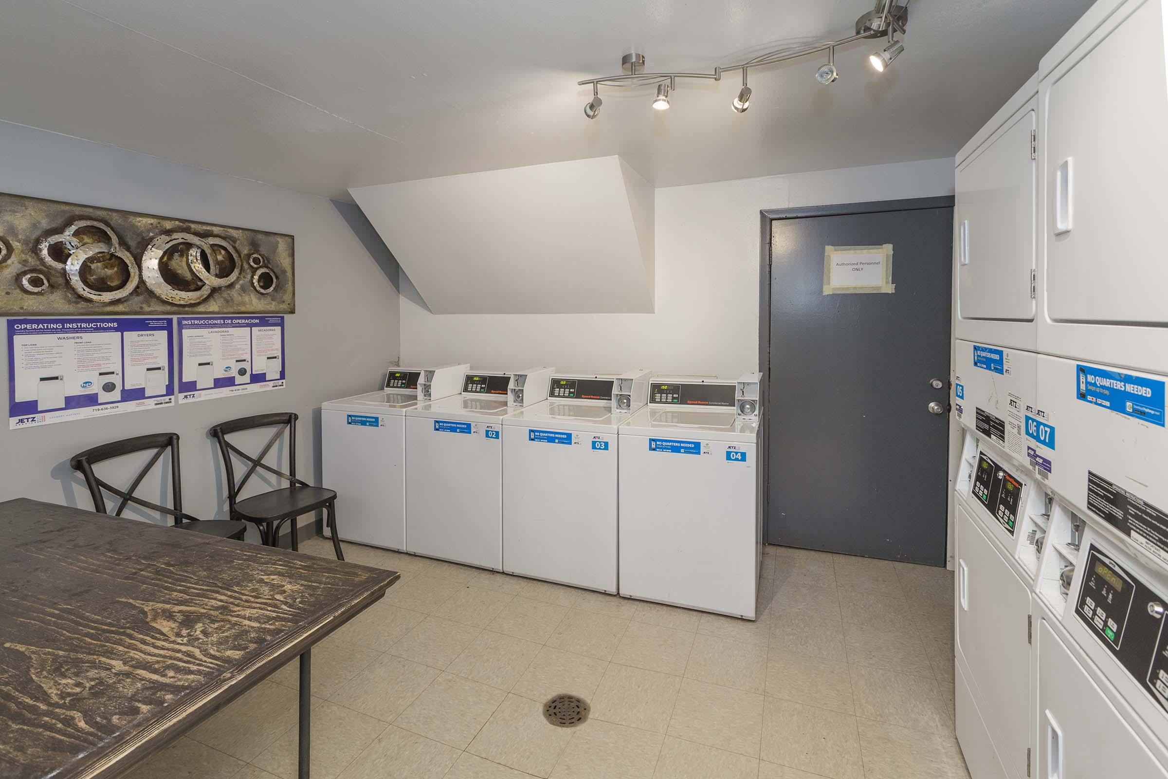 A laundry room featuring several white washing machines and dryers lined up against the wall. There's a table with two chairs, and a door with a sign can be seen. The walls are painted in light colors, and a piece of abstract art is displayed on one wall. The floor is tiled.