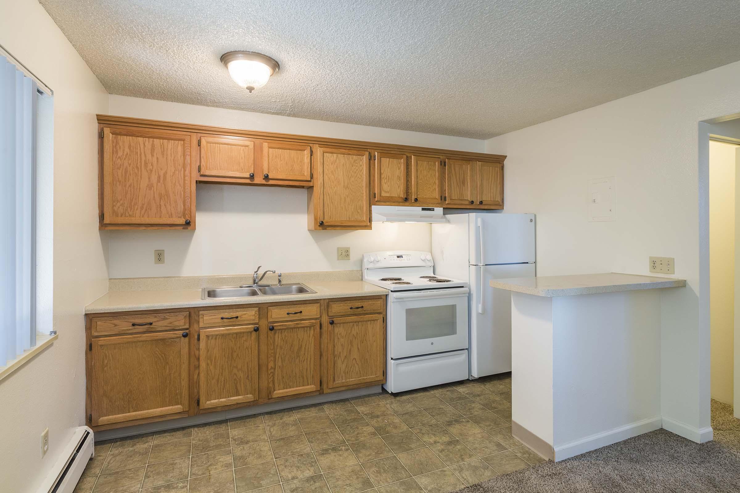 A kitchen with wooden cabinets, a countertop, and essential appliances including a sink, stove, oven, and refrigerator. The space features neutral walls and a light-colored floor, creating a bright and airy atmosphere. A small bar area is visible, providing additional counter space.