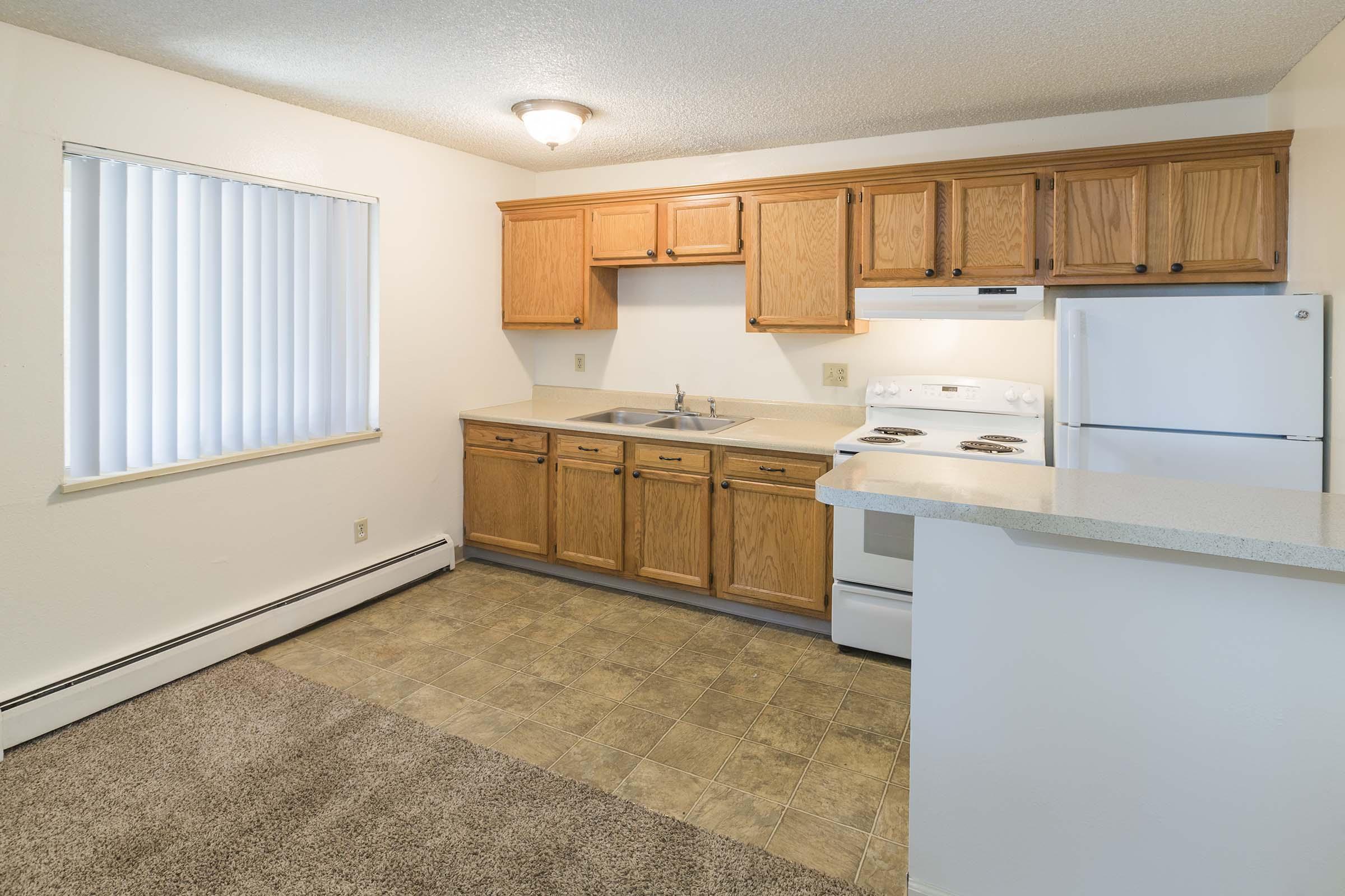 A small kitchen featuring light wood cabinets, a white stove, oven, and refrigerator. The counter is beige with a sink, and there's a window with vertical blinds. The floor is covered with vinyl tiles, and a carpeted area is visible in the foreground. The overall color scheme is neutral and bright.