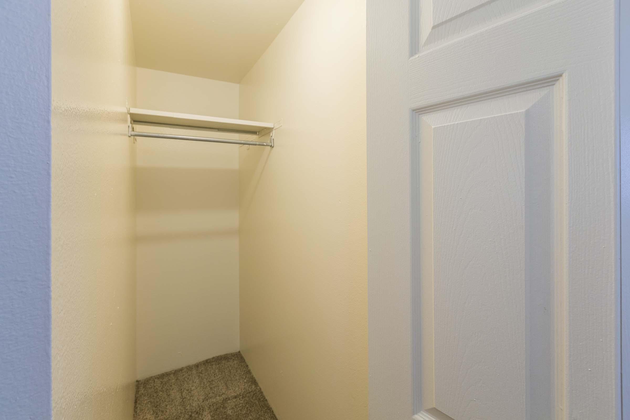 An empty closet with a hanging rod and beige walls, featuring a carpeted floor. The door is partially visible on the right, and the space appears clean and unoccupied.
