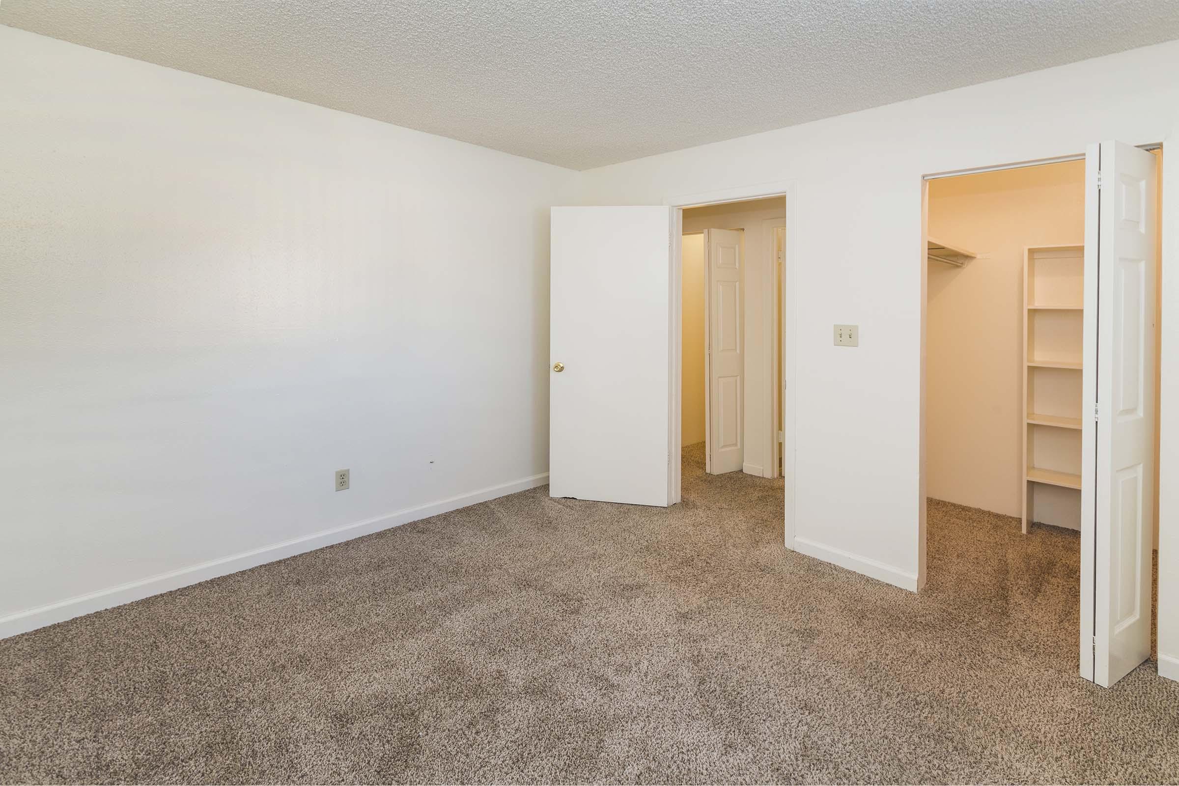 A light and airy room with beige carpet, featuring a white-painted wall. Two open doors reveal a closet and a small adjoining space. Natural light enters the room, creating a welcoming atmosphere. The overall look is clean and simple, suitable for a bedroom or personal space.