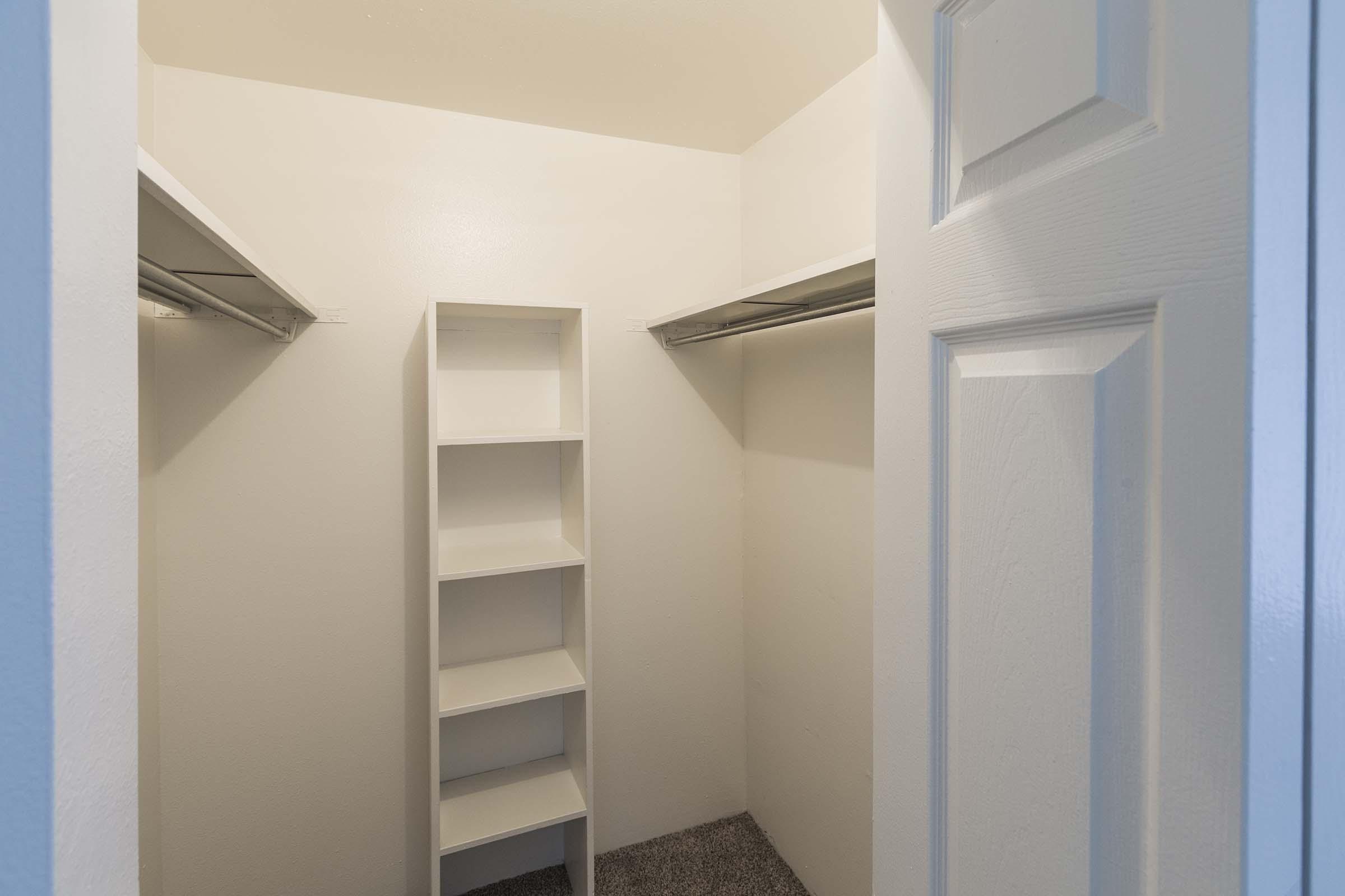 A view of a minimalist walk-in closet featuring two clothing racks on either side and a narrow white shelving unit in the center. The walls are plain and painted white, and the floor is covered in soft gray carpet. The closet door is partially open, revealing its spacious interior.