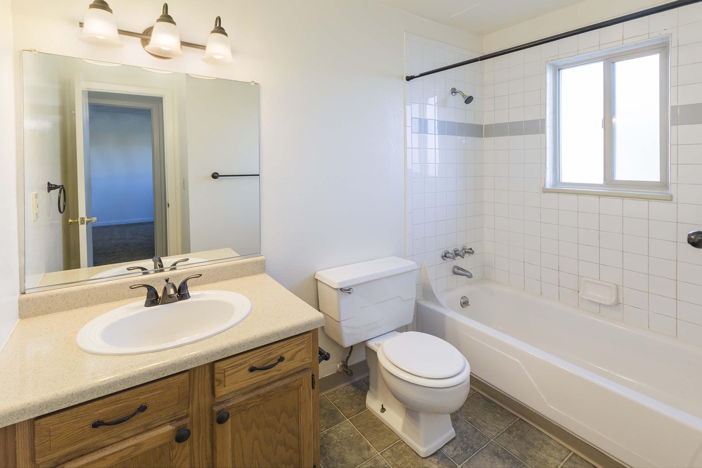 A clean, well-lit bathroom featuring a bathtub with a shower, a toilet, and a sink with a wooden cabinet underneath. The walls are tiled with white and gray accents, and there is a large mirror above the sink. A window lets in natural light, and the floor is tiled in a gray pattern.