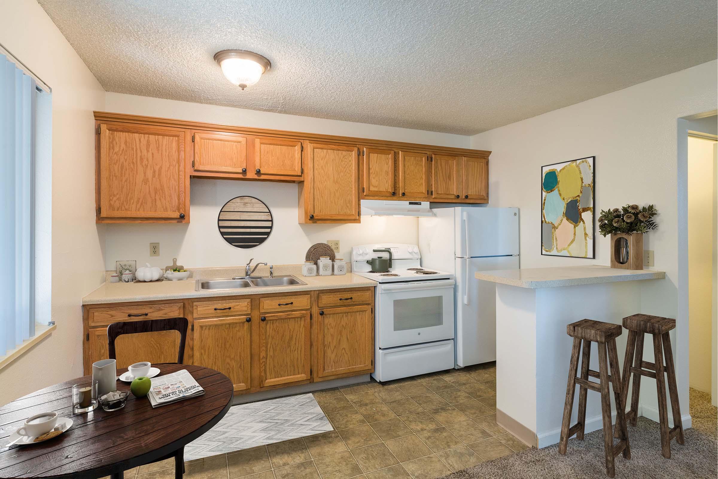 Cozy kitchen featuring wooden cabinetry, a sink, stove, and refrigerator. A small dining table is set with dishes, and there are two high stools at a breakfast bar. The room has neutral-colored walls and a circular mirror on the wall. Natural light is coming through a window, enhancing the inviting atmosphere.