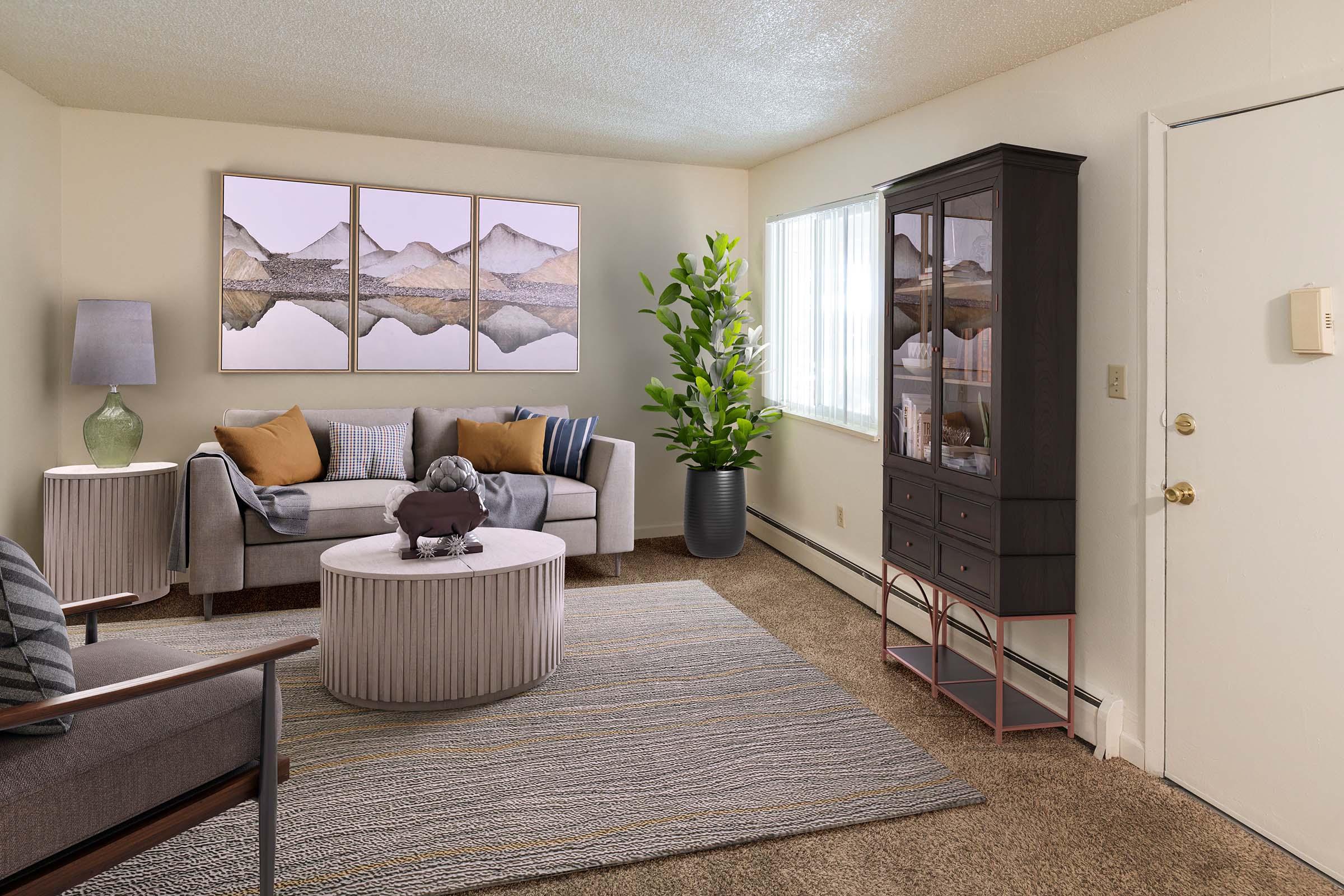A cozy living room featuring a light-colored sofa with patterned cushions, a round coffee table, and a cabinet displaying decorative items. A large green plant adds a touch of nature, while three framed mountain landscape pictures adorn the wall. The space is brightly lit by a window with sheer curtains.