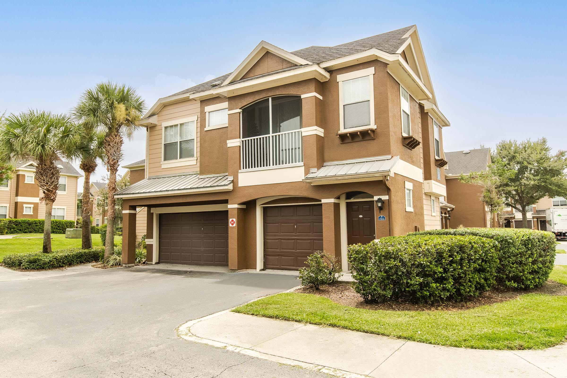 Two-story residential building with beige and brown exterior, featuring a balcony on the second floor. The ground level has two garage doors, and the property is surrounded by well-maintained grass and palm trees. Clear blue sky in the background complements the scene.