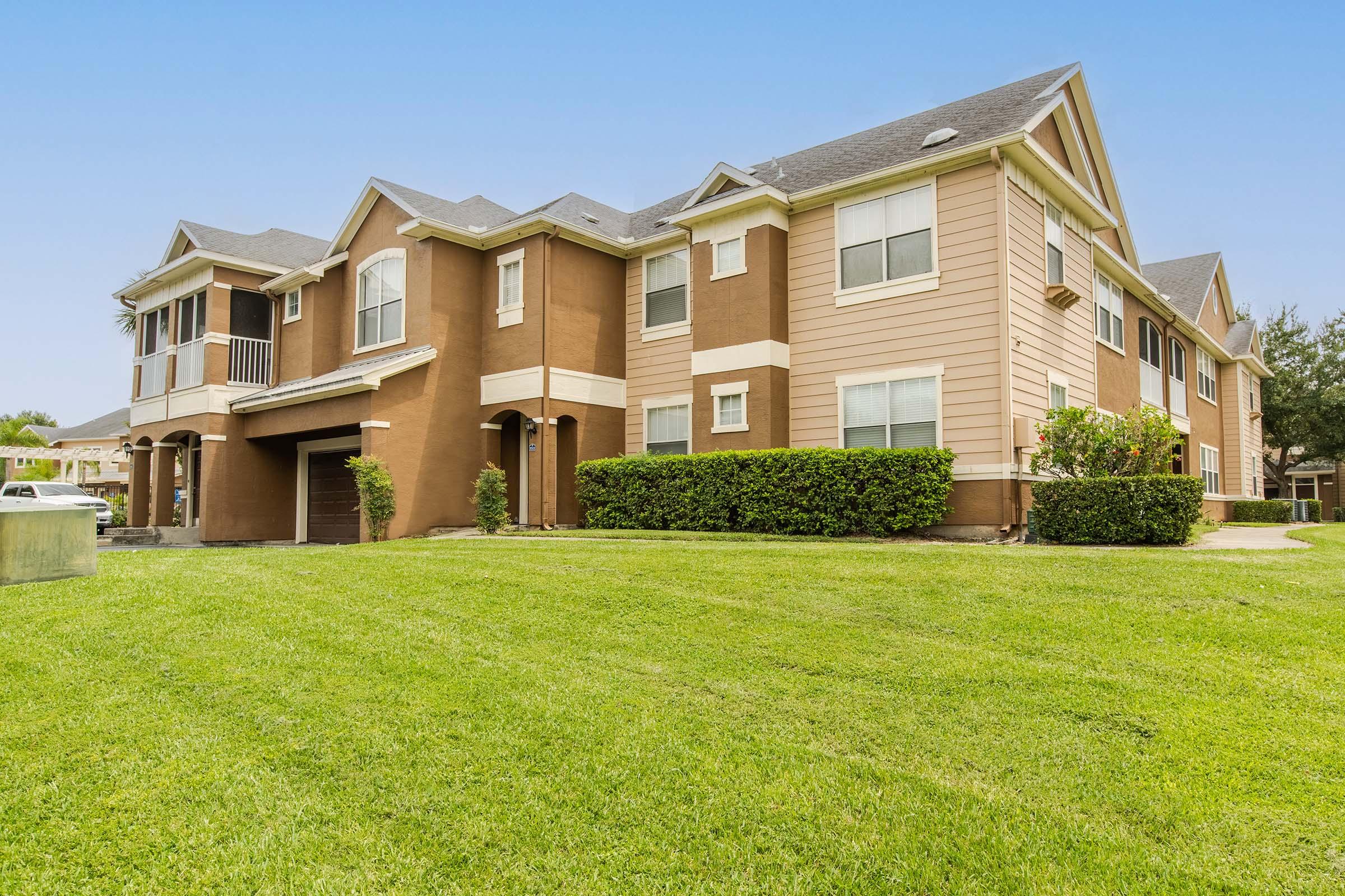 A residential building featuring a mix of architectural styles, with beige and brown siding. The property includes a manicured lawn and shrubs in front, with several windows and a garage visible. The sky is clear, suggesting a sunny day.