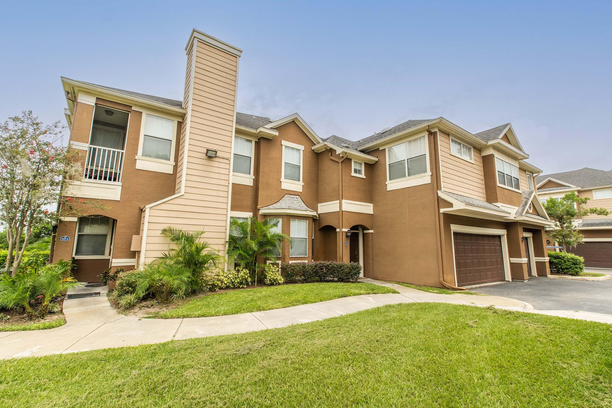A two-story residential building with a brown exterior and multiple balconies. A well-maintained lawn with green grass surrounds the property, and there are shrubs and small trees in the landscaping. An attached garage is visible on the right side of the building. Clear blue sky in the background.