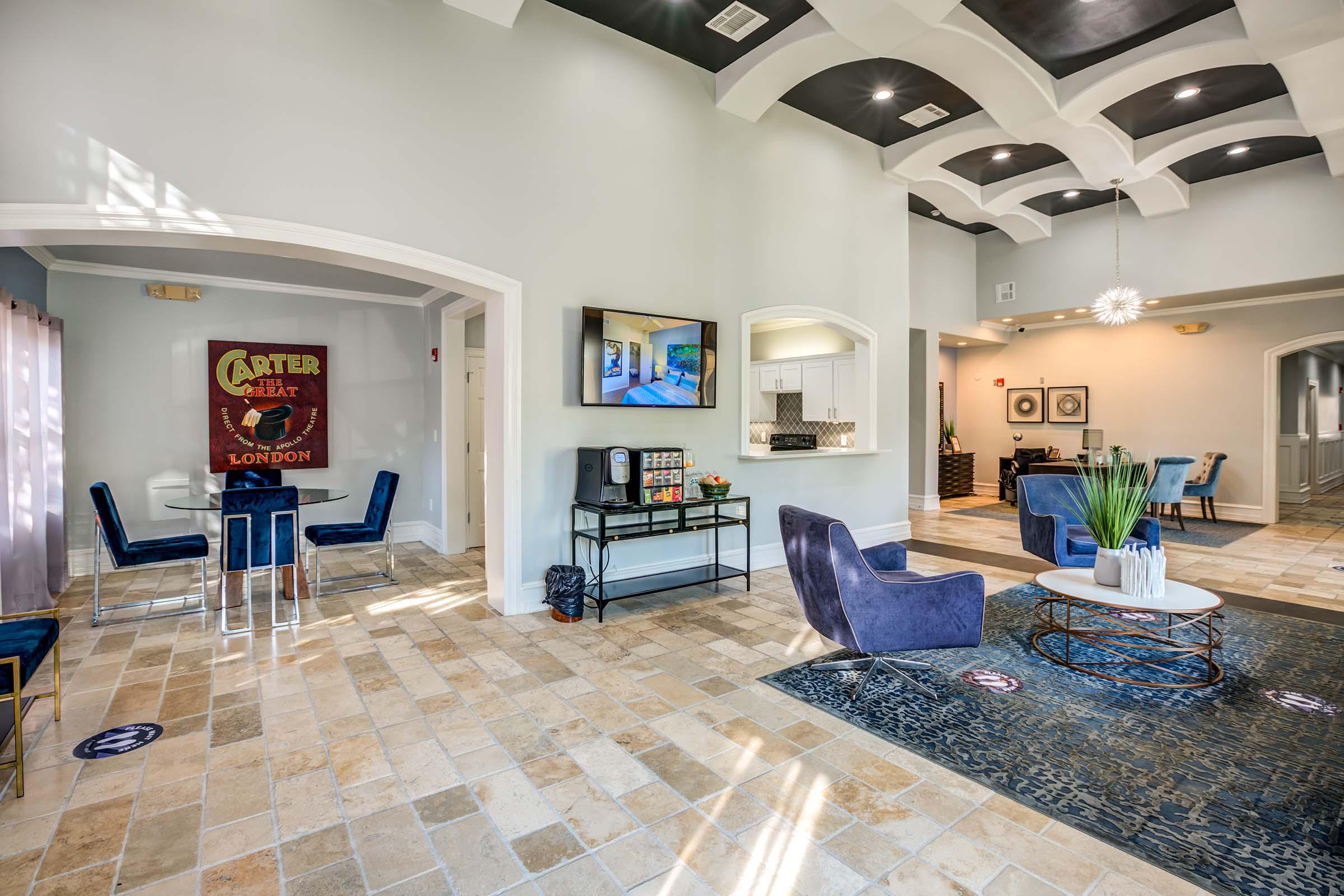 Interior view of a modern lobby featuring arched ceilings and a mix of blue and beige furnishings. A seating area with stylish chairs and a coffee table is visible, along with a wall-mounted TV and a kitchenette. Decorative elements include a large rug and plants, creating a welcoming atmosphere.
