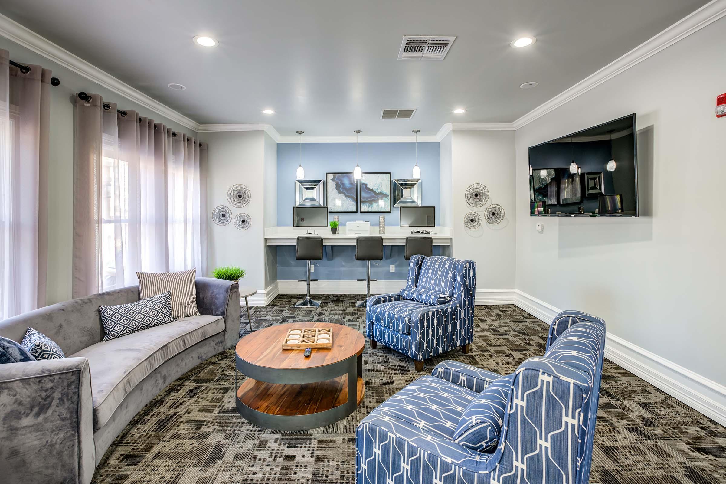 Cozy living area featuring a grey sofa and two blue patterned chairs around a round coffee table. A small bar area with high stools is visible in the background, along with a large wall-mounted TV. Natural light streams in through the windows, enhancing the modern decor and inviting atmosphere.