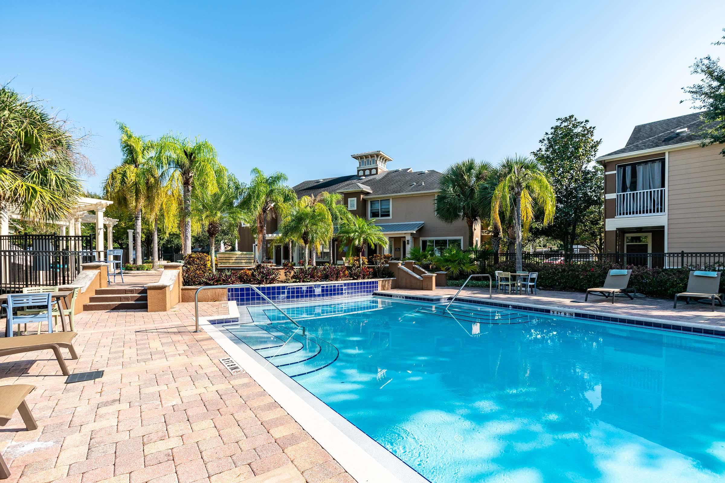 A sunny outdoor pool area surrounded by palm trees and well-maintained landscaping. Lounge chairs are set around the pool, and there are several buildings in the background, suggesting an apartment complex. The clear blue sky enhances the inviting atmosphere of the space.