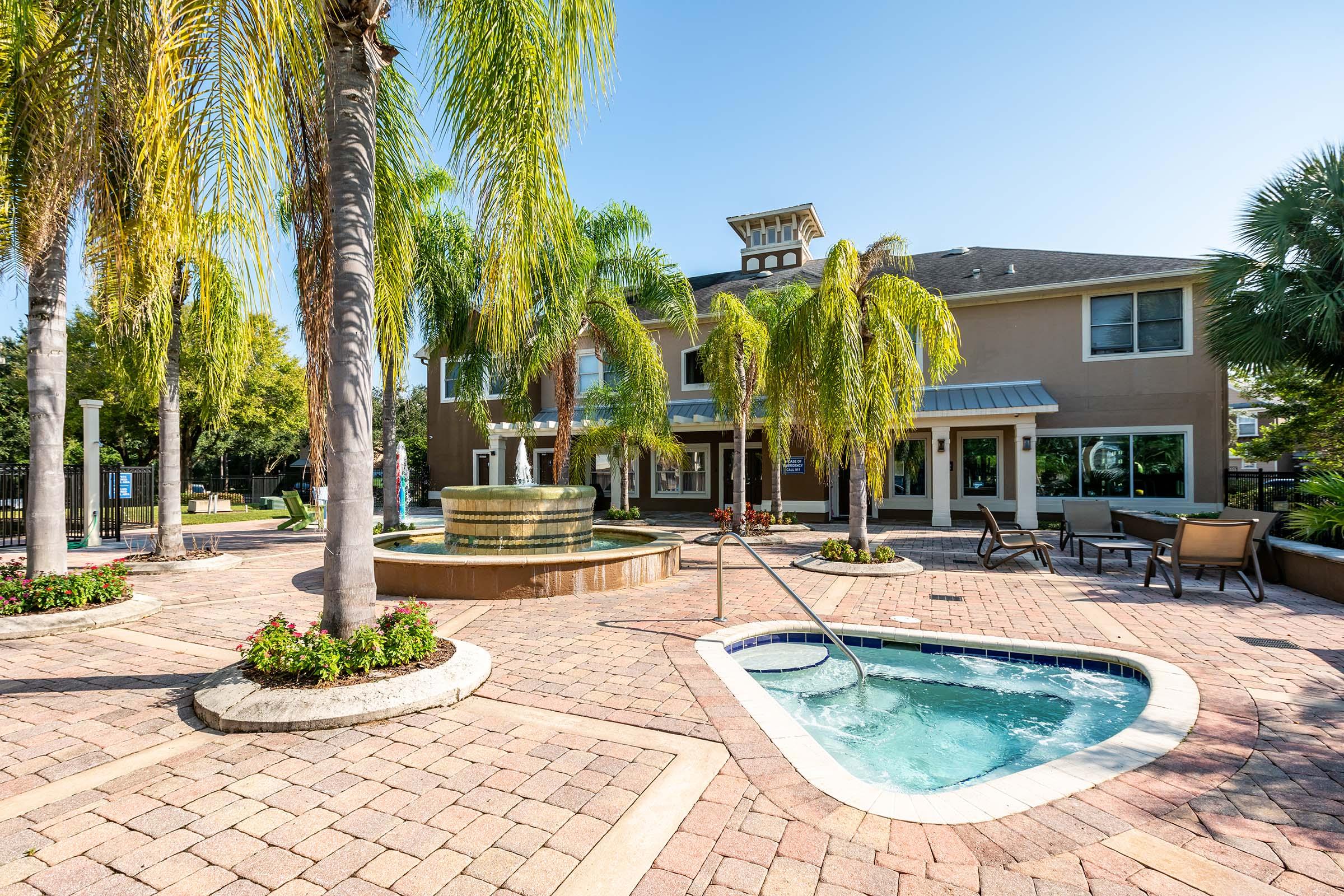 A landscaped pool area featuring a hot tub and a fountain surrounded by palm trees. In the background, a modern building with large windows and seating areas. The setting is bright and inviting, suggesting a resort-like atmosphere.