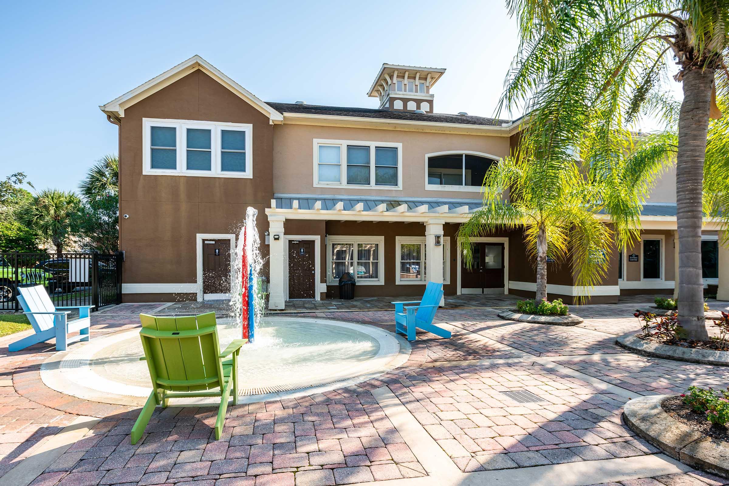 A modern building with a brown facade, featuring large windows. In front, there’s a small splash pad with colorful water features and a seating area with blue and green chairs. Palm trees are visible, and the surrounding area is paved with bricks, creating a welcoming outdoor space.