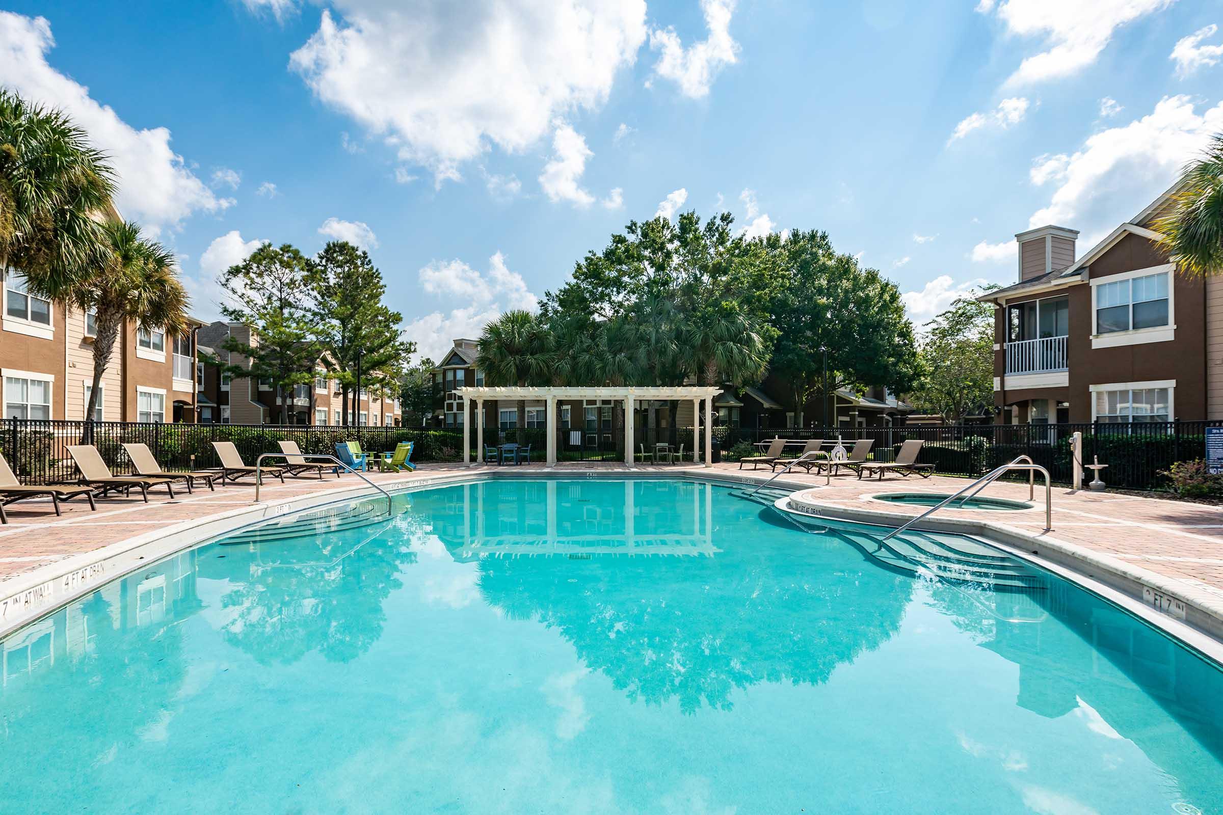 A clear blue swimming pool surrounded by lounge chairs and palm trees, with a shaded seating area in the background. Bright blue sky with fluffy clouds above creates a serene and inviting atmosphere.