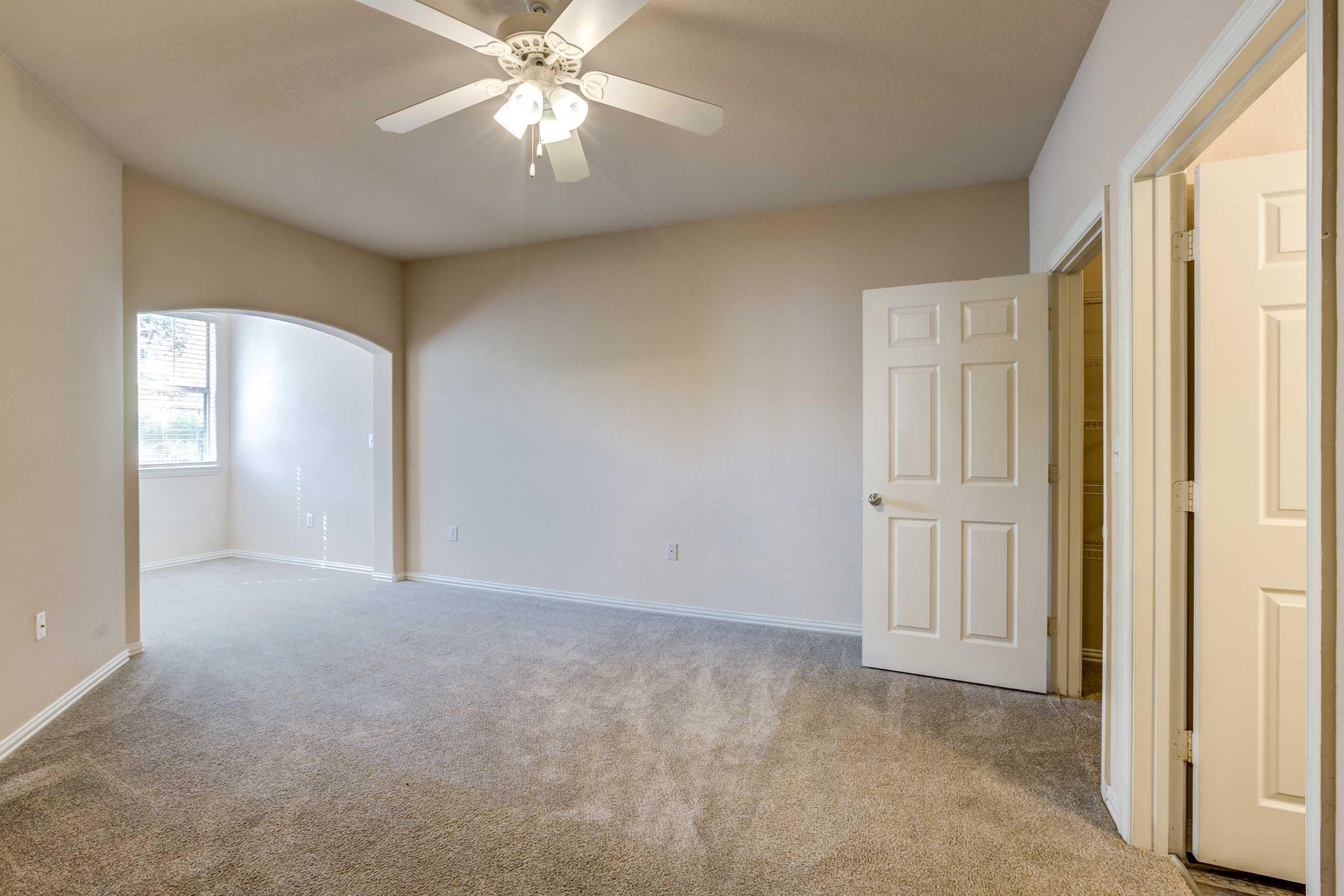 A spacious, empty room with beige walls and carpet. A ceiling fan is mounted above, and there are two doorways leading to other areas. Natural light enters through a window, and the room features a modern, clean design, making it suitable for various uses.