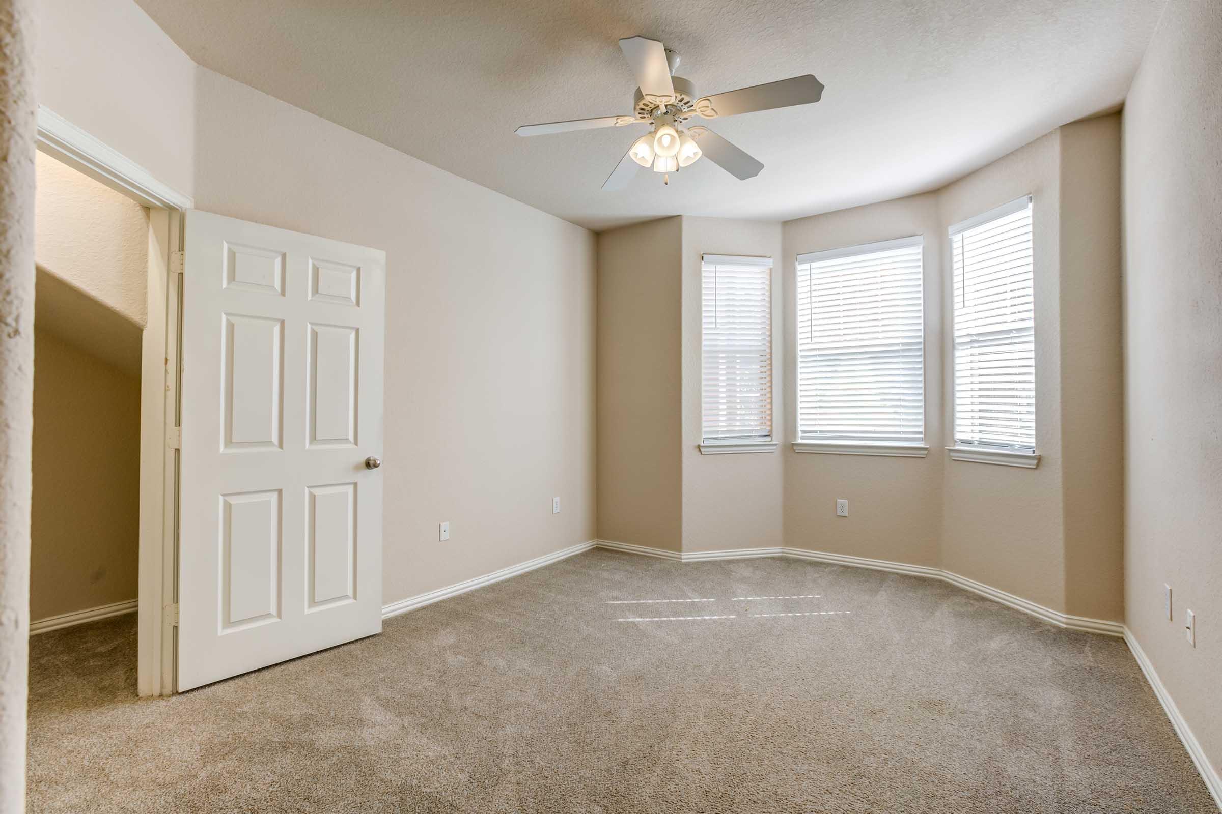 Empty room with light beige walls and carpet, featuring a ceiling fan. There are three windows with blinds allowing natural light, and a closed white door on the left leading to another space. The overall atmosphere is bright and clean, suitable for various uses.