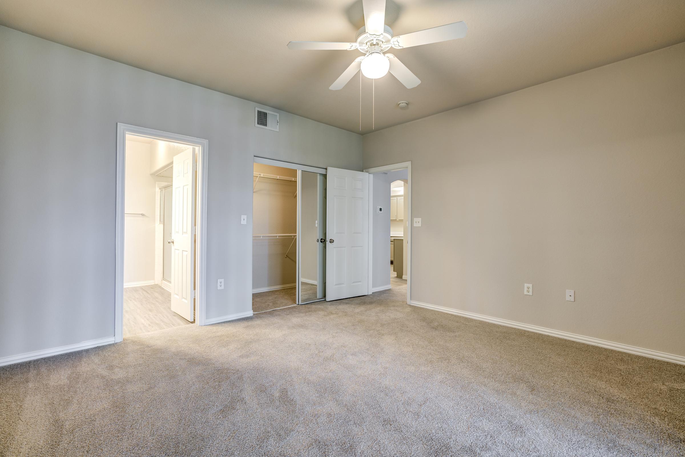 A spacious, well-lit room with light gray walls and plush carpet. It features a ceiling fan, two doors leading to a closet and bathroom, and ample space for furniture. The overall atmosphere is inviting and modern, ideal for a bedroom or living area.