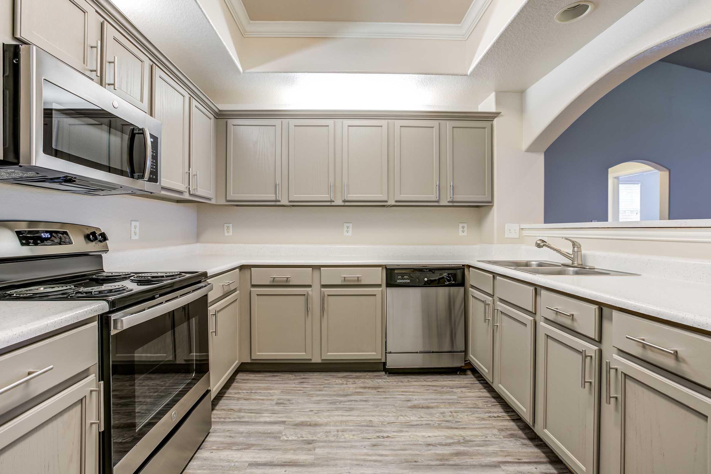 Modern kitchen featuring gray cabinets, stainless steel appliances, and a combination of a gas stove and dishwasher. The countertop is light-colored, and the flooring has a laminate wood finish. The space is well-lit with multiple light fixtures and an open layout.