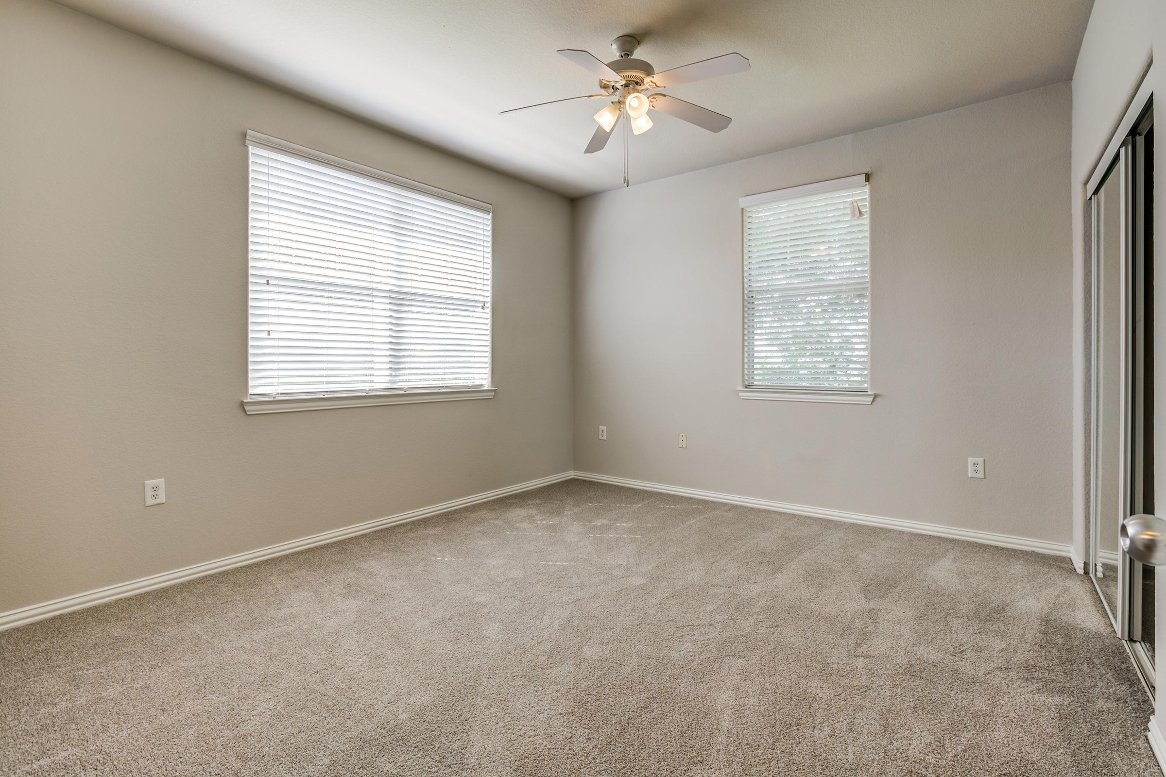 A spacious, empty room with light gray walls and beige carpet. It features two windows with white blinds allowing natural light, a ceiling fan, and minimal decor. There's a closet to the right with sliding doors, creating a clean and inviting atmosphere.