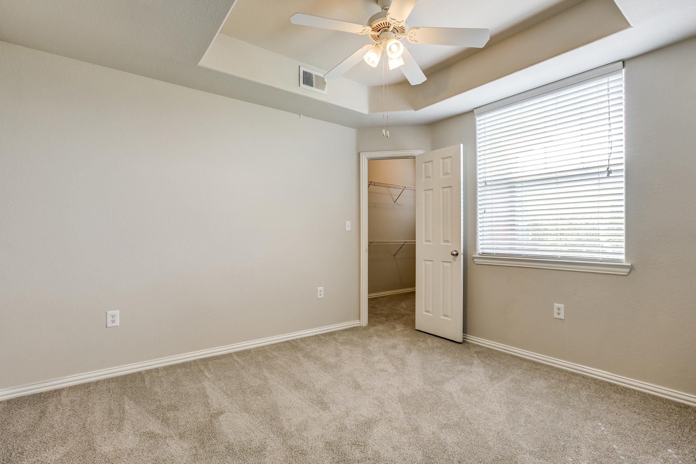 A spacious, well-lit empty bedroom featuring light gray walls, carpeting, a ceiling fan, and a window with blinds. A door leads to a small closet, and the room is designed with a neutral color palette, enhancing its airy ambiance. Ideal for personalization and decorating.