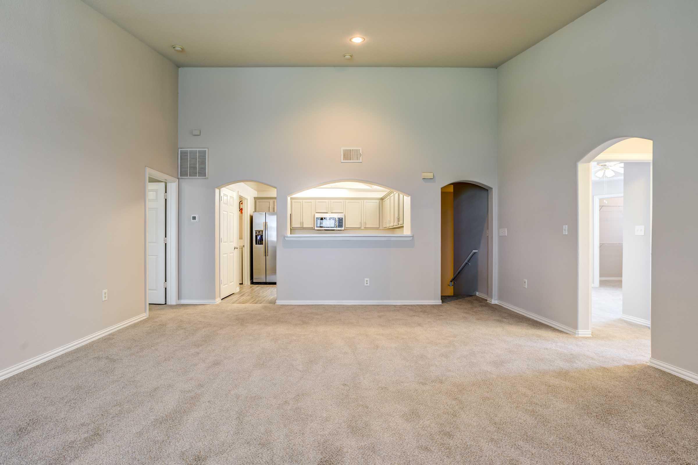 Spacious, light-filled living room with neutral walls and soft carpet flooring. Two doorways lead to adjacent areas, one with a staircase and another to a kitchen with light-colored cabinetry visible in the background. The room features a high ceiling and natural light from windows.