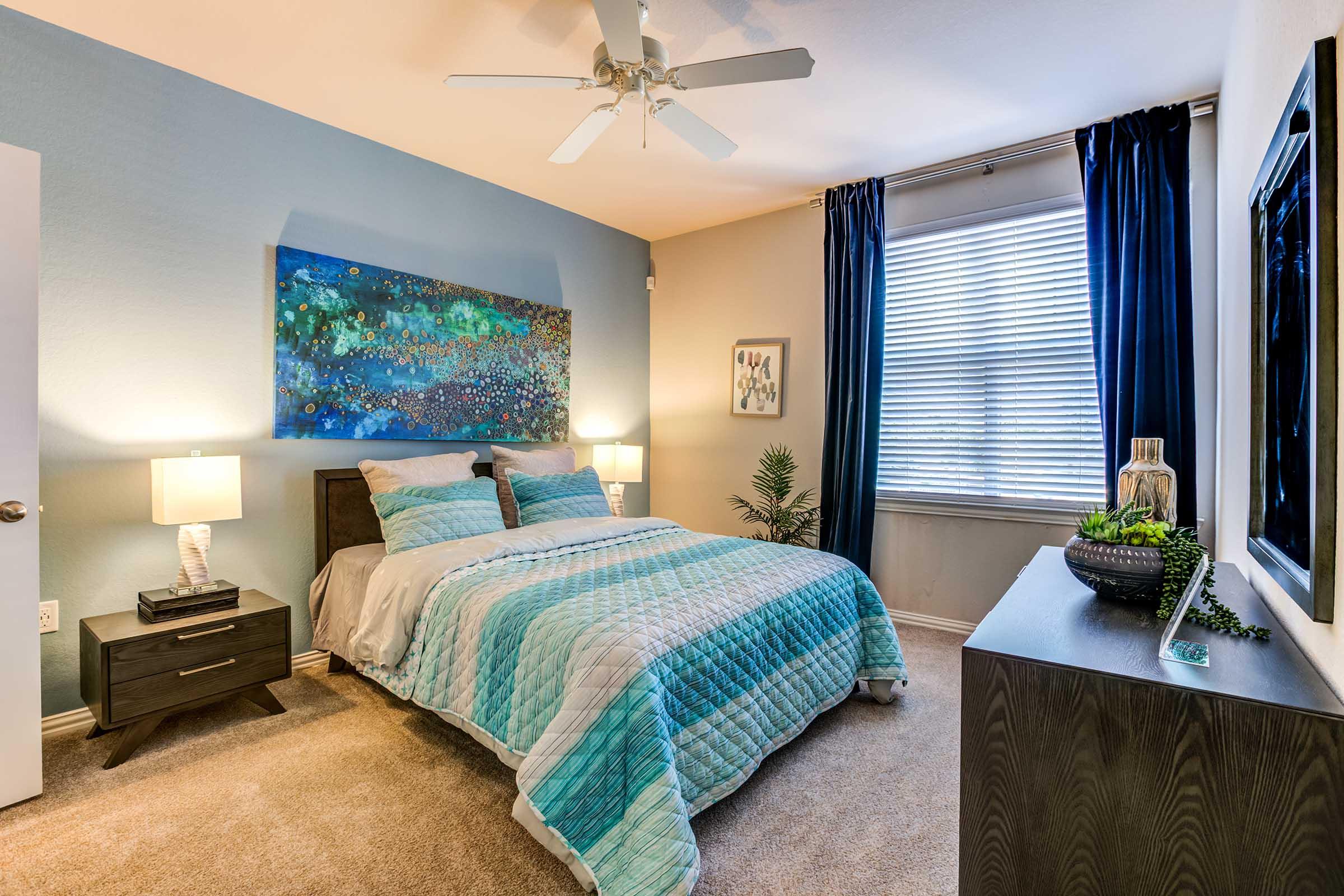 A cozy bedroom featuring a queen-sized bed with a blue and white patterned quilt, two bedside lamps, a large window with blue curtains, and a decorative painting on the wall. The room has light beige walls, a small indoor plant, and a dark wooden dresser. Natural light brightens the space.
