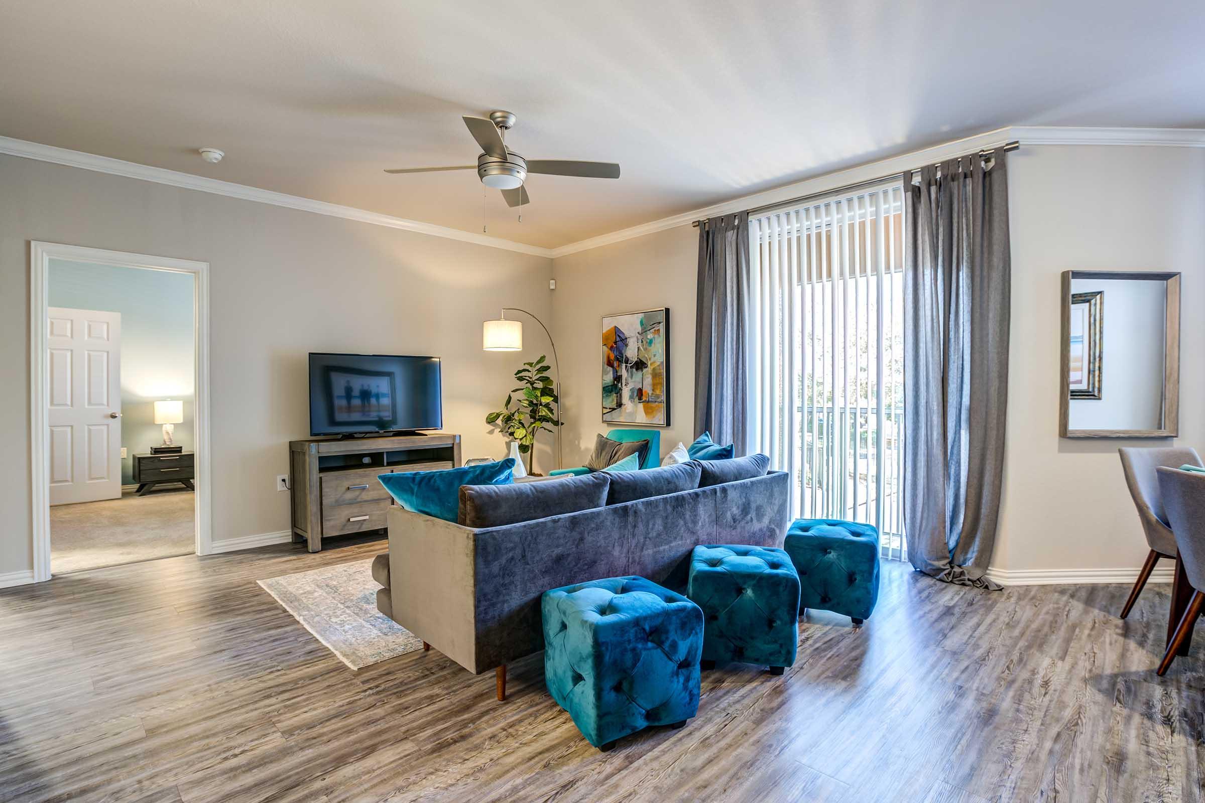 A modern living room featuring a gray sofa with teal accent stools, a TV on a wooden cabinet, and large windows with sheer curtains. The walls are painted in light colors, complemented by a piece of artwork and a floor lamp. Natural light fills the space, creating a welcoming atmosphere.
