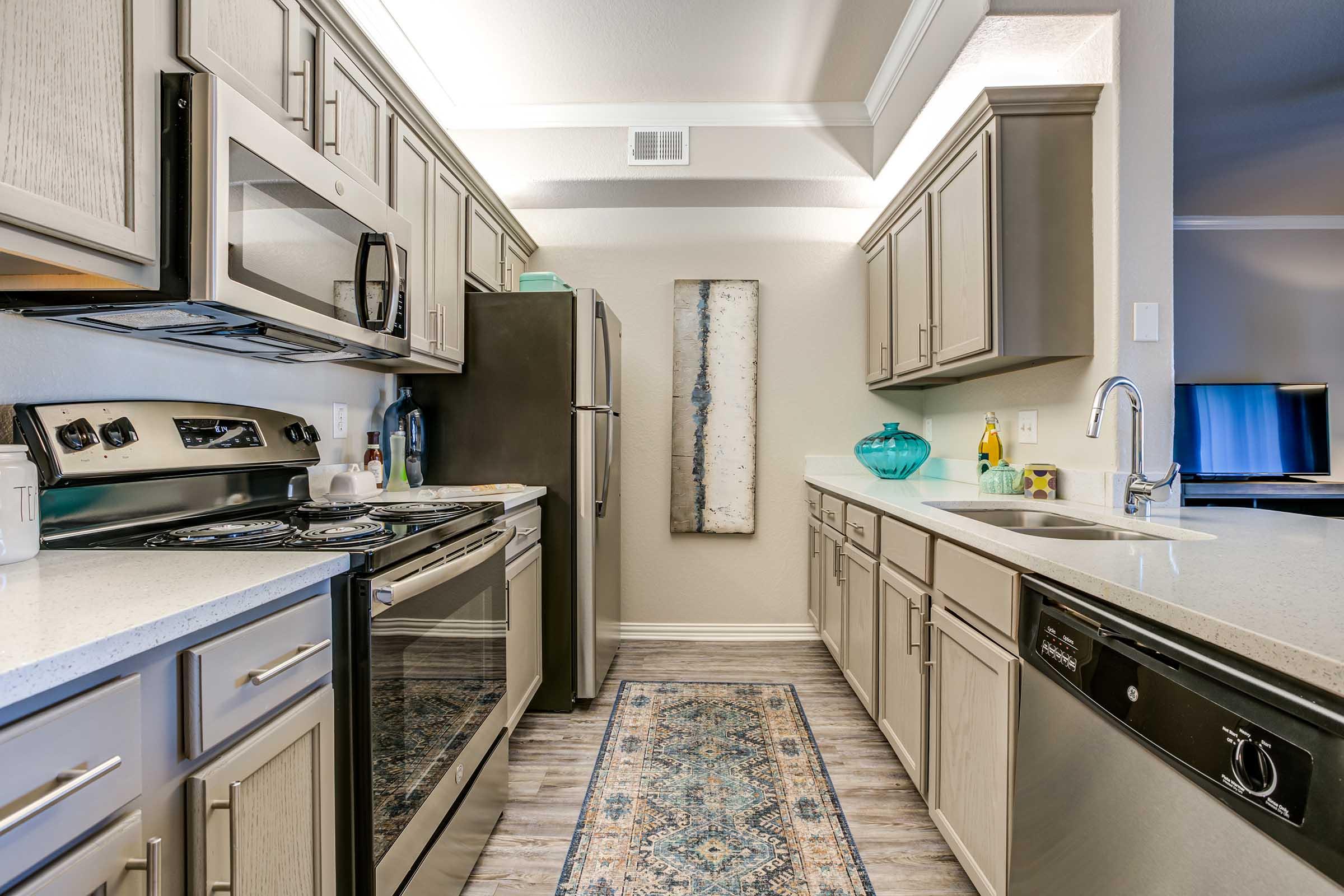 Modern kitchen featuring stainless steel appliances, including a stove, microwave, and refrigerator. The cabinetry is light gray, complemented by a light-colored countertop. A decorative rug in the center adds color, while a wall art piece and decorative items enhance the aesthetic. Natural light fills the space.