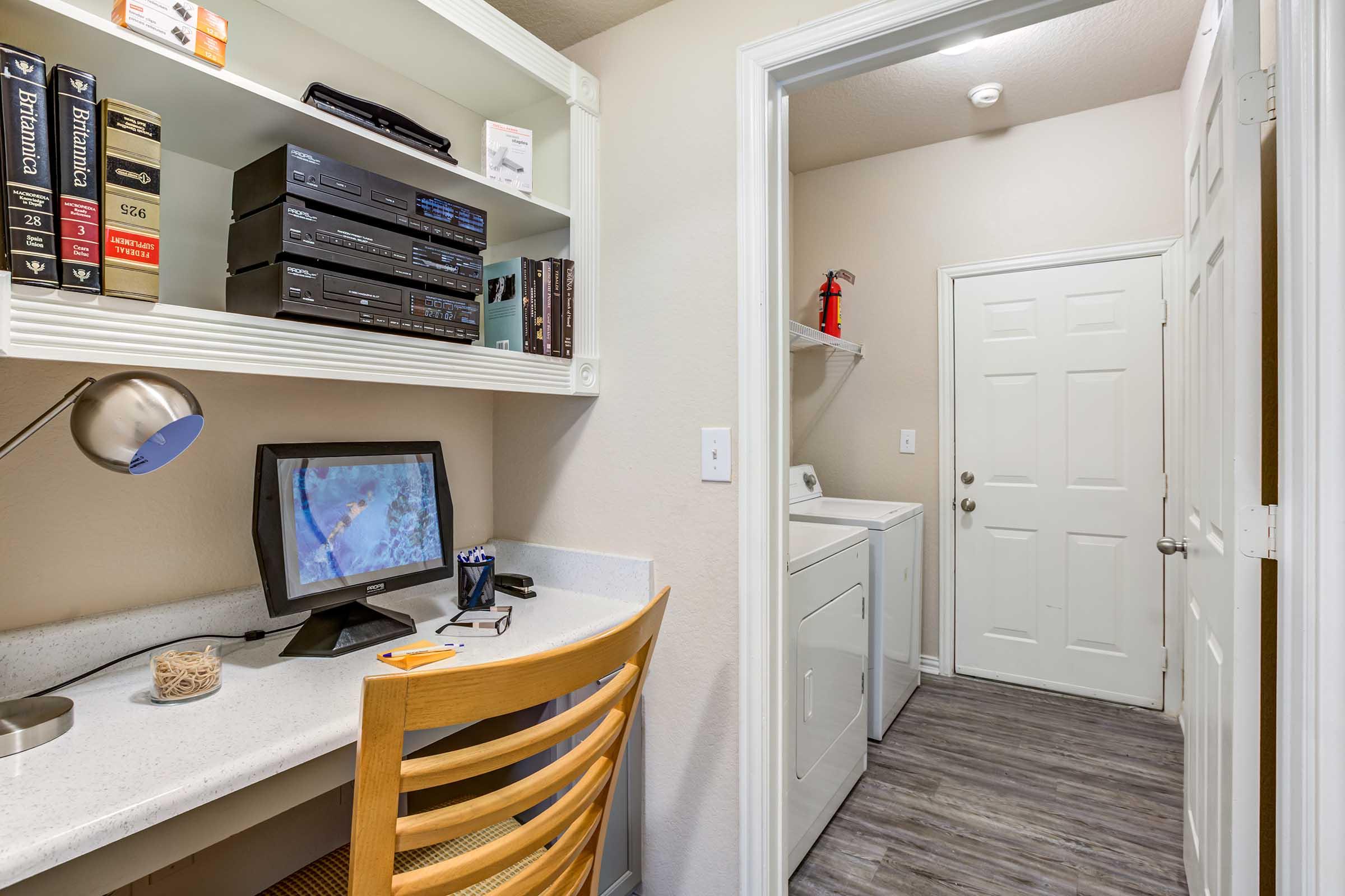 A small home office space featuring a desk with a monitor and a lamp, wooden chair, and various books on shelves above. To the right, there's a doorway leading to a laundry area with a washing machine and a fire extinguisher mounted on the wall. The walls are painted light colors, creating a bright atmosphere.