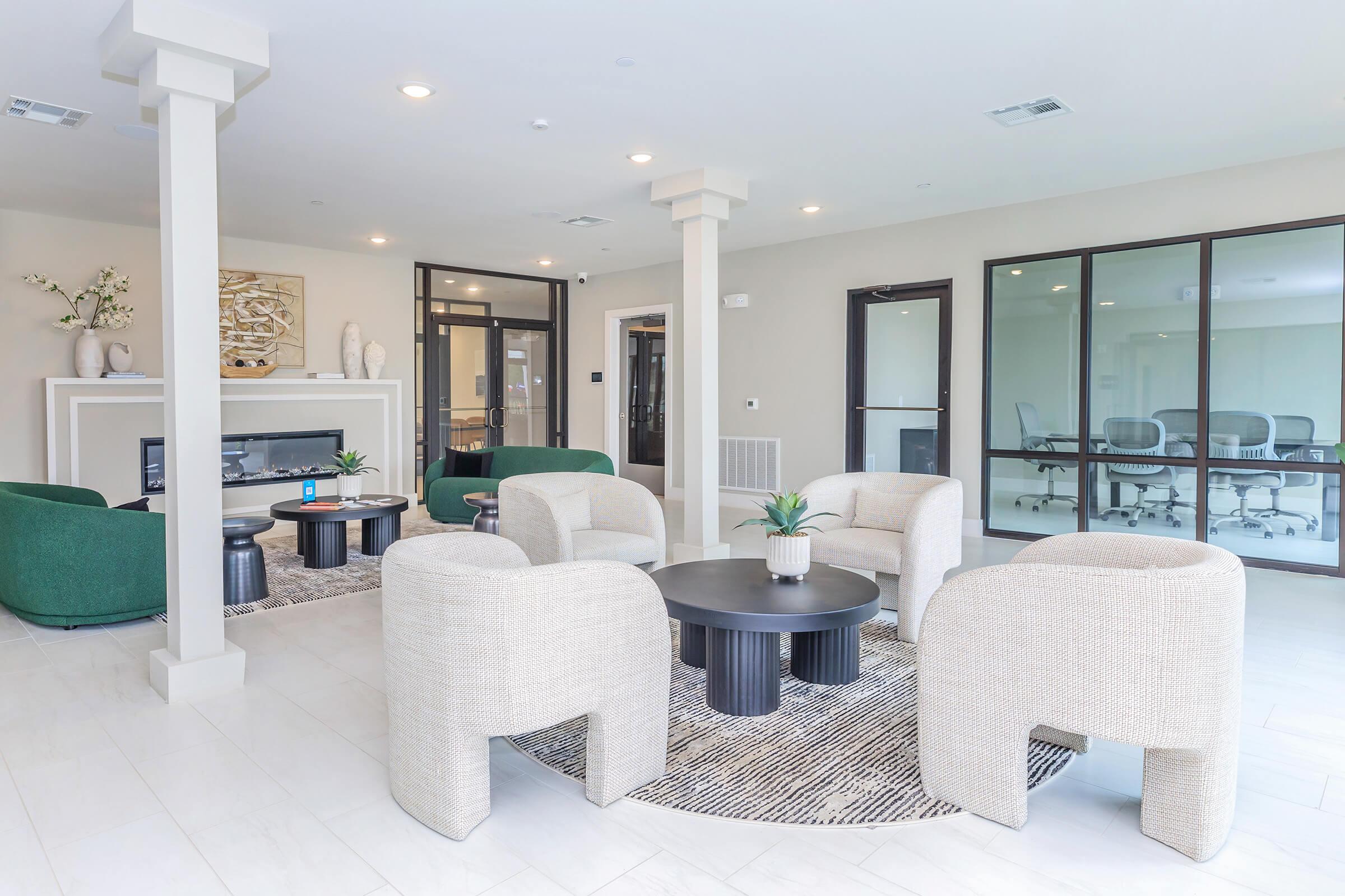 A modern lounge area featuring light-colored walls and elegant furnishings. There are two green armchairs, circular coffee tables, and several small decorative items. Large glass doors lead to a conference room, enhancing the open and welcoming atmosphere.