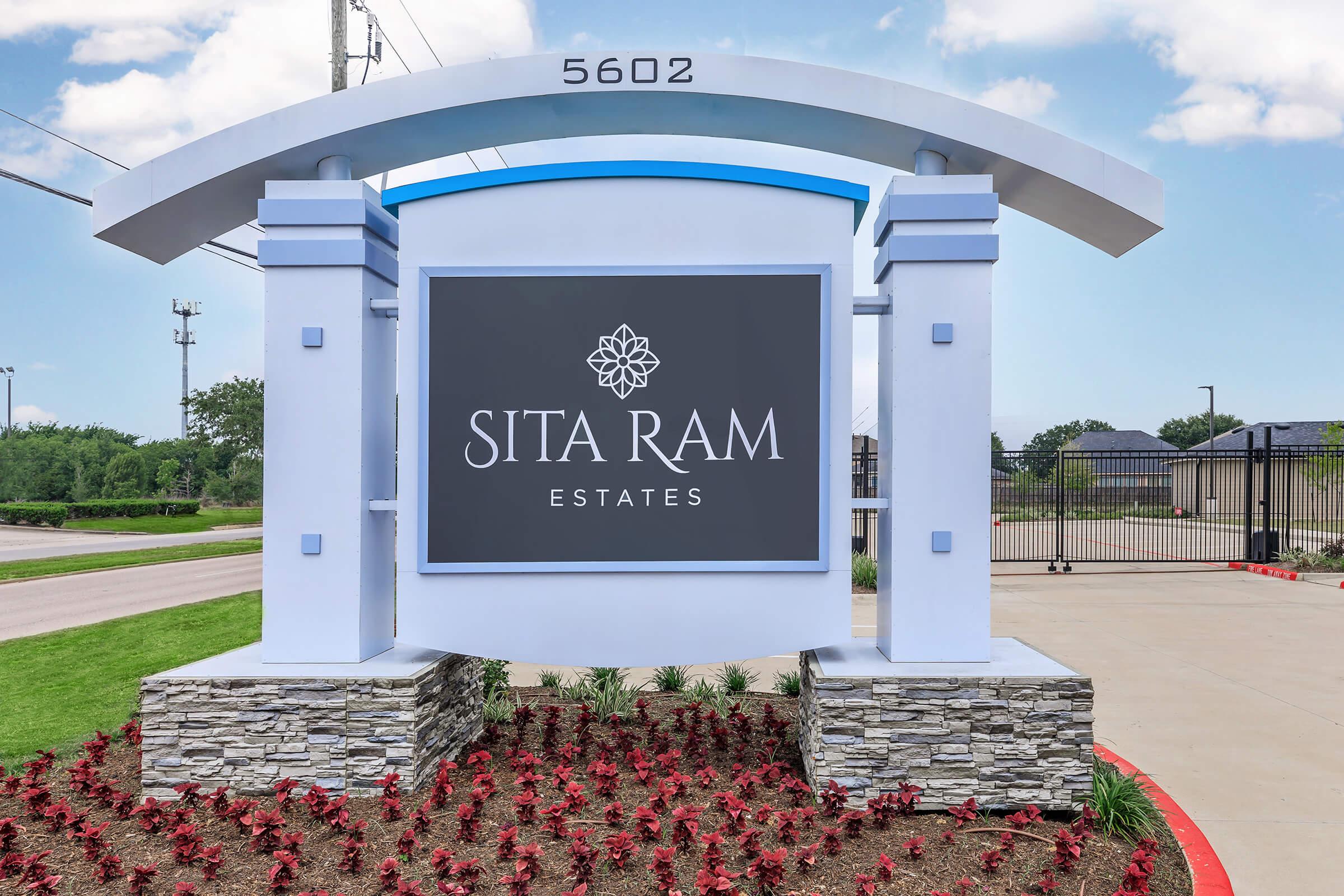 Sign for Sita Ram Estates featuring bold lettering on a dark background. The sign is set in front of a landscaped area with red flowers and stone accents. It is situated on a property entrance, with a clear blue sky in the background. The street number displayed is 5602.