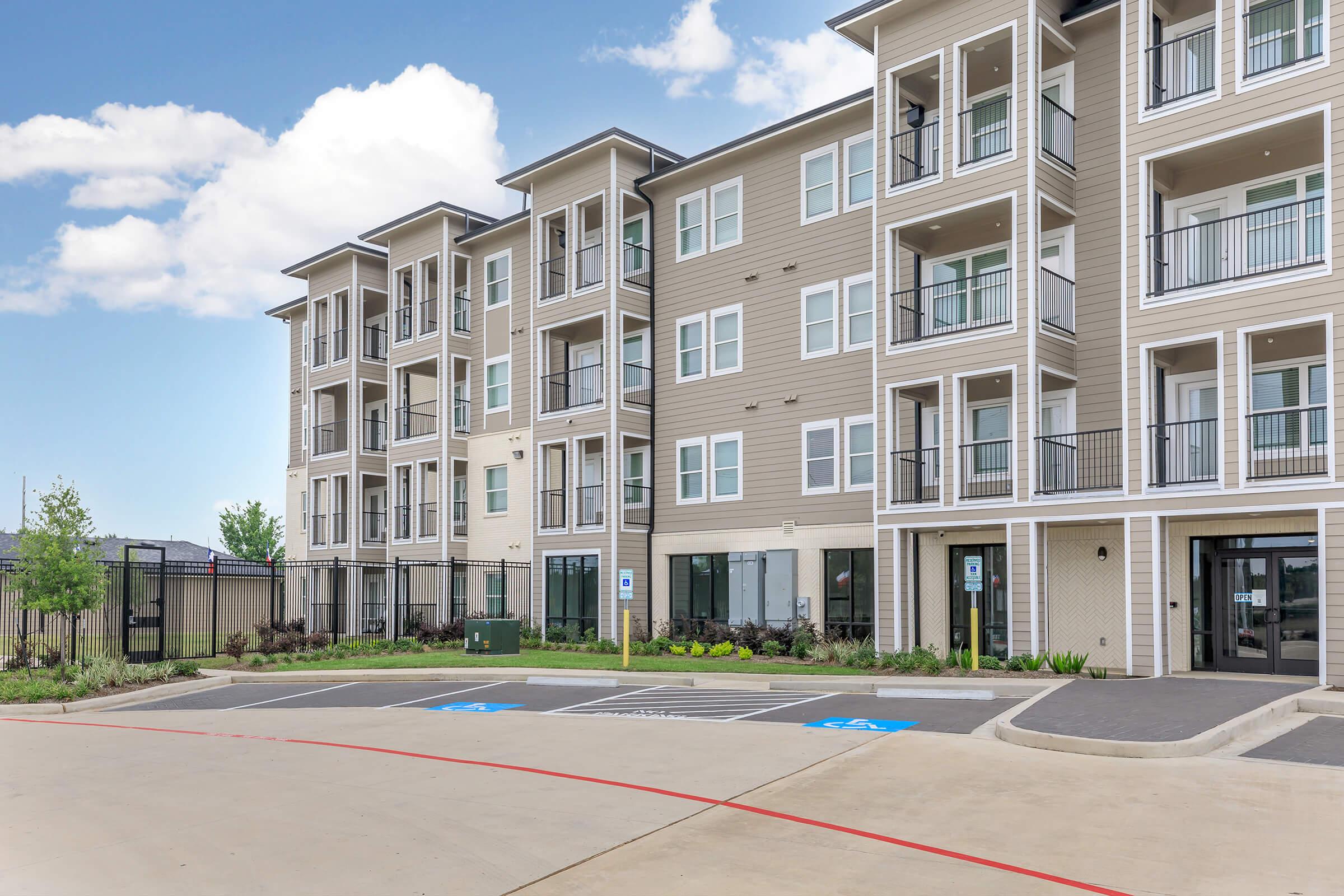 Exterior view of a modern, multi-story apartment building featuring a mix of balconies, large windows, and a driveway. The structure has beige siding and is surrounded by landscaped areas, including trees and shrubs. Blue skies with clouds are visible in the background.