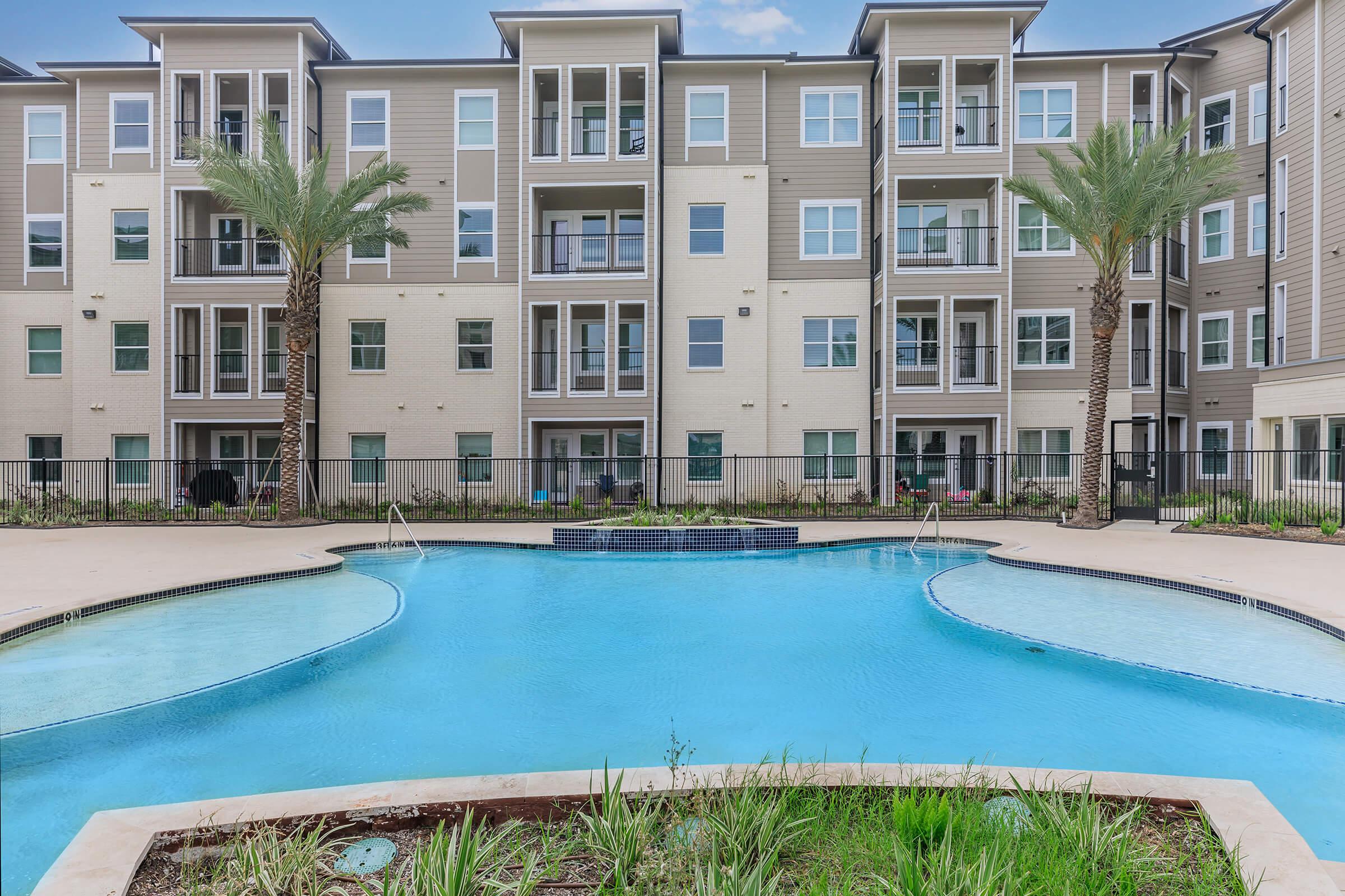 A modern apartment complex featuring multiple stories with balconies, surrounded by palm trees. In the foreground, a blue swimming pool with a curved edge, set in a landscaped area with grass and plants. The building has a contemporary design with large windows.