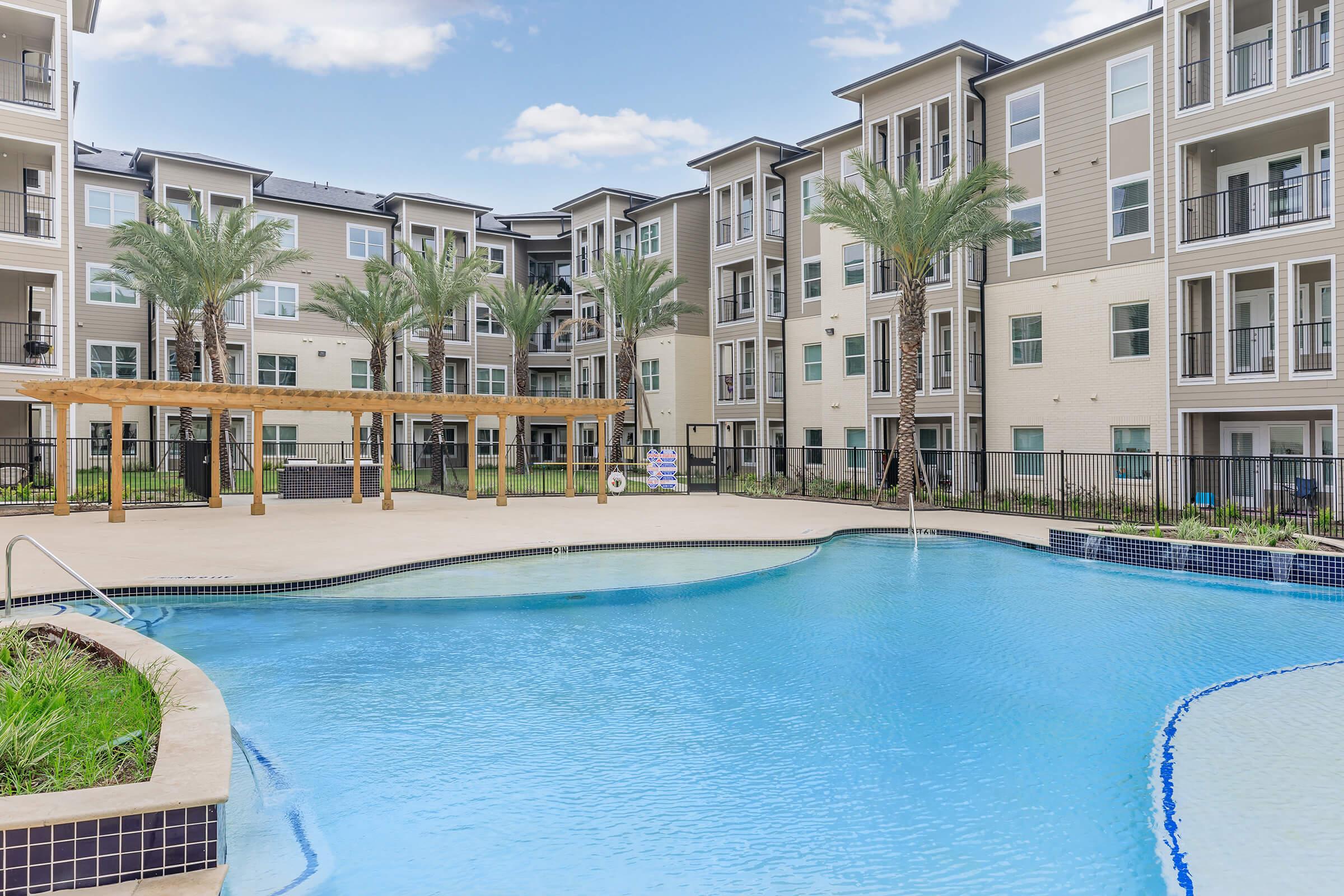 A spacious outdoor pool surrounded by palm trees, with modern apartment buildings in the background. The pool area features a wooden pergola and lounge space, creating an inviting atmosphere for relaxation. The sky is clear with a few clouds.