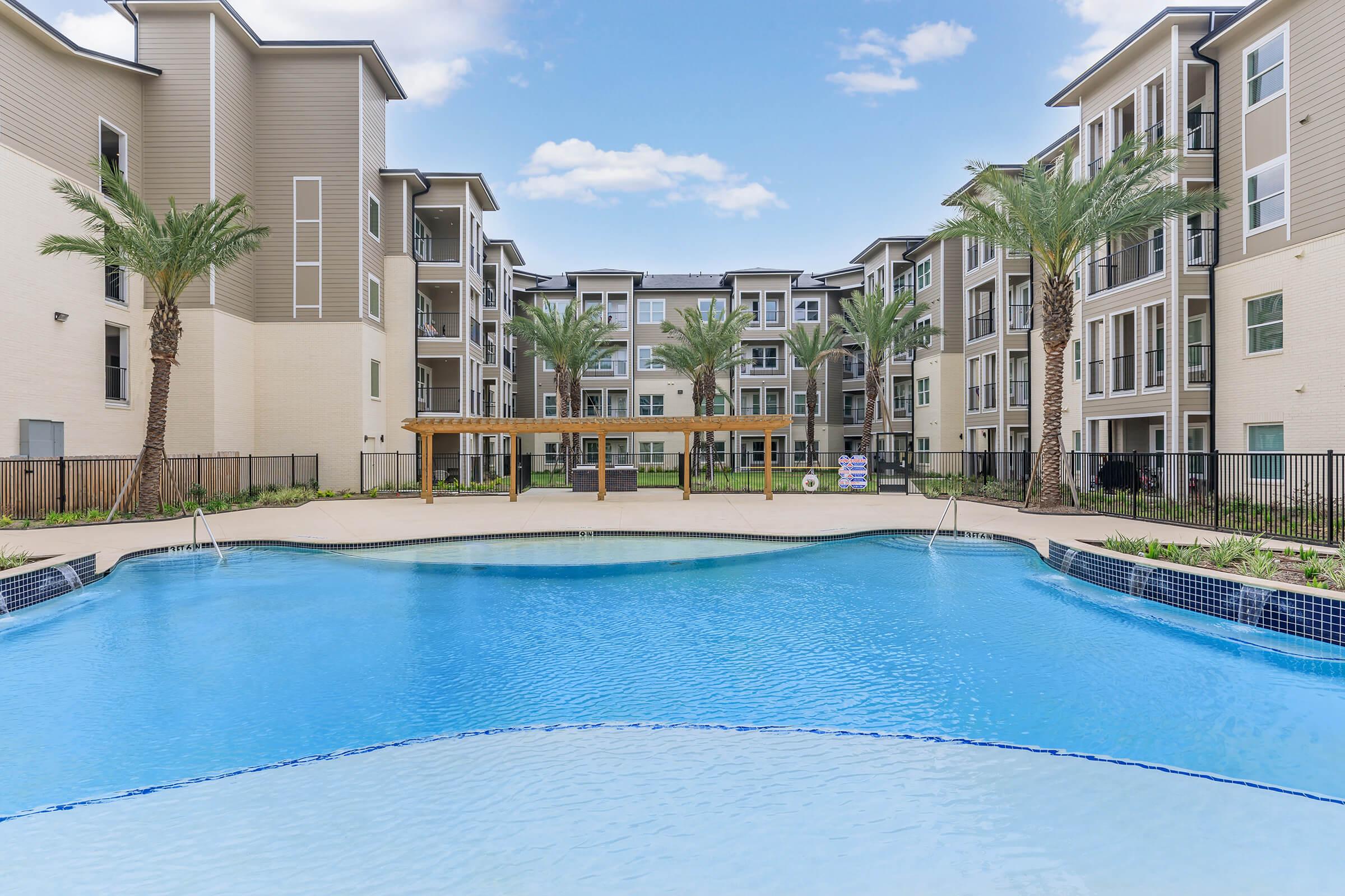 A sparkling swimming pool surrounded by modern apartment buildings and palm trees, with a wooden pergola and landscaped greenery in the courtyard area. The sky is clear, creating a bright and inviting atmosphere.