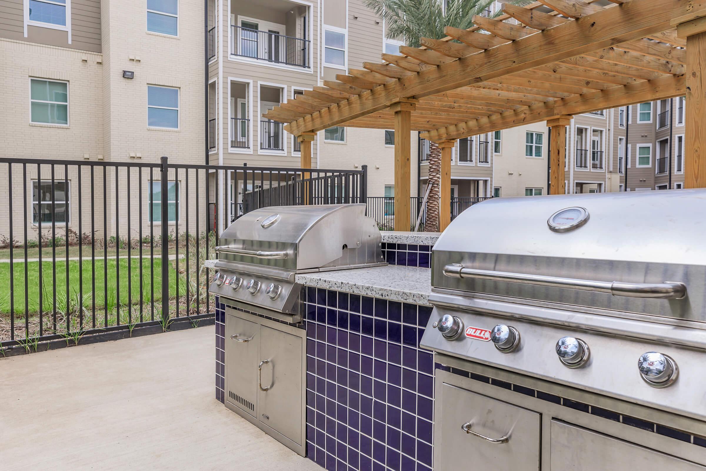 Outdoor barbecue area featuring two stainless steel grills with granite countertops, surrounded by a fenced patio and landscaped greenery. In the background, a multi-story apartment building adds to the setting. The space is designed for communal cooking and social gatherings.