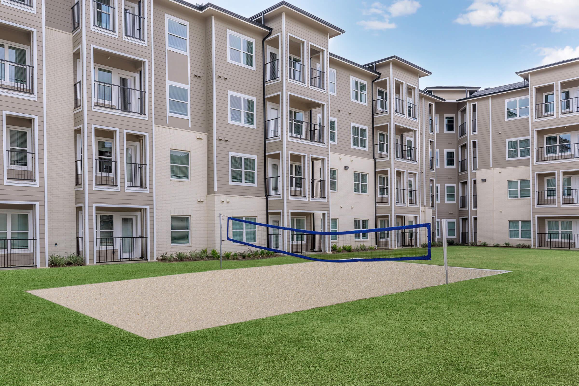 An outdoor volleyball court located in a grassy area between modern apartment buildings, featuring a sand-filled court with boundary lines and a net set up for play. The surrounding apartments are three stories tall with balconies and large windows, creating a spacious and inviting atmosphere.
